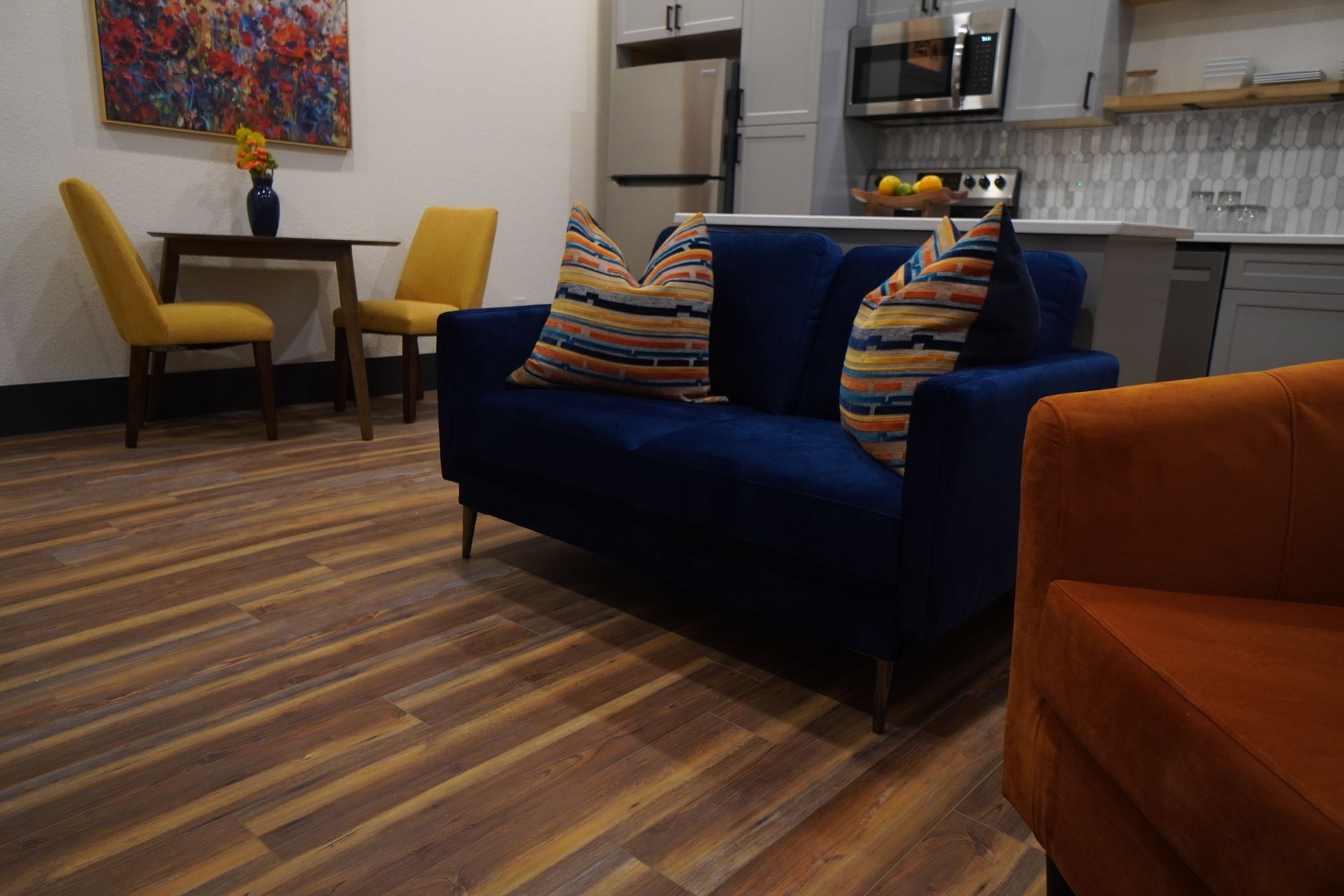 Living room with blue loveseat, orange armchair, yellow chairs, and kitchen in the background. Wooden floors.