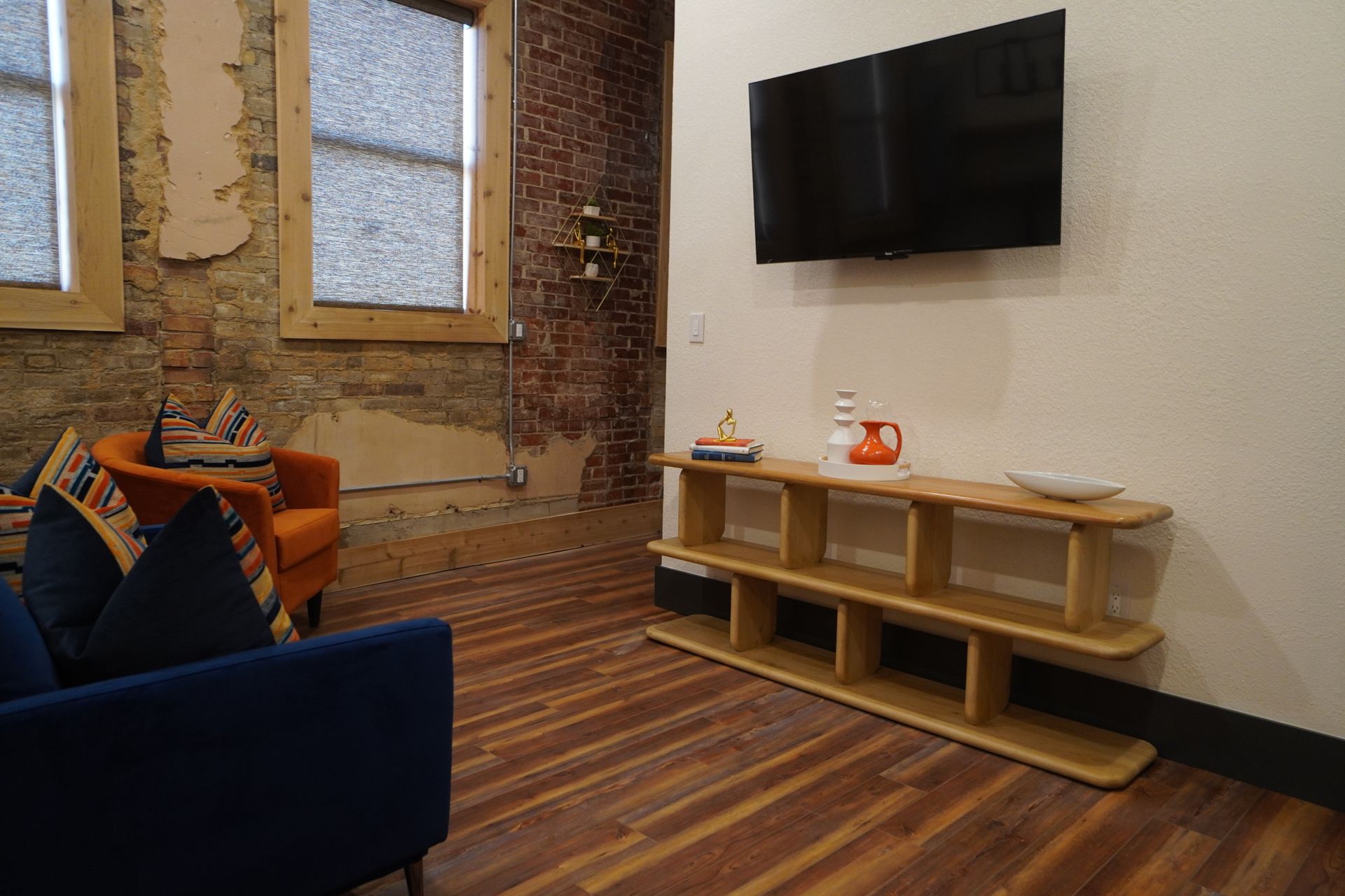 Living room with exposed brick wall, wood floor, and blue sofa. Orange chair and TV on wall.