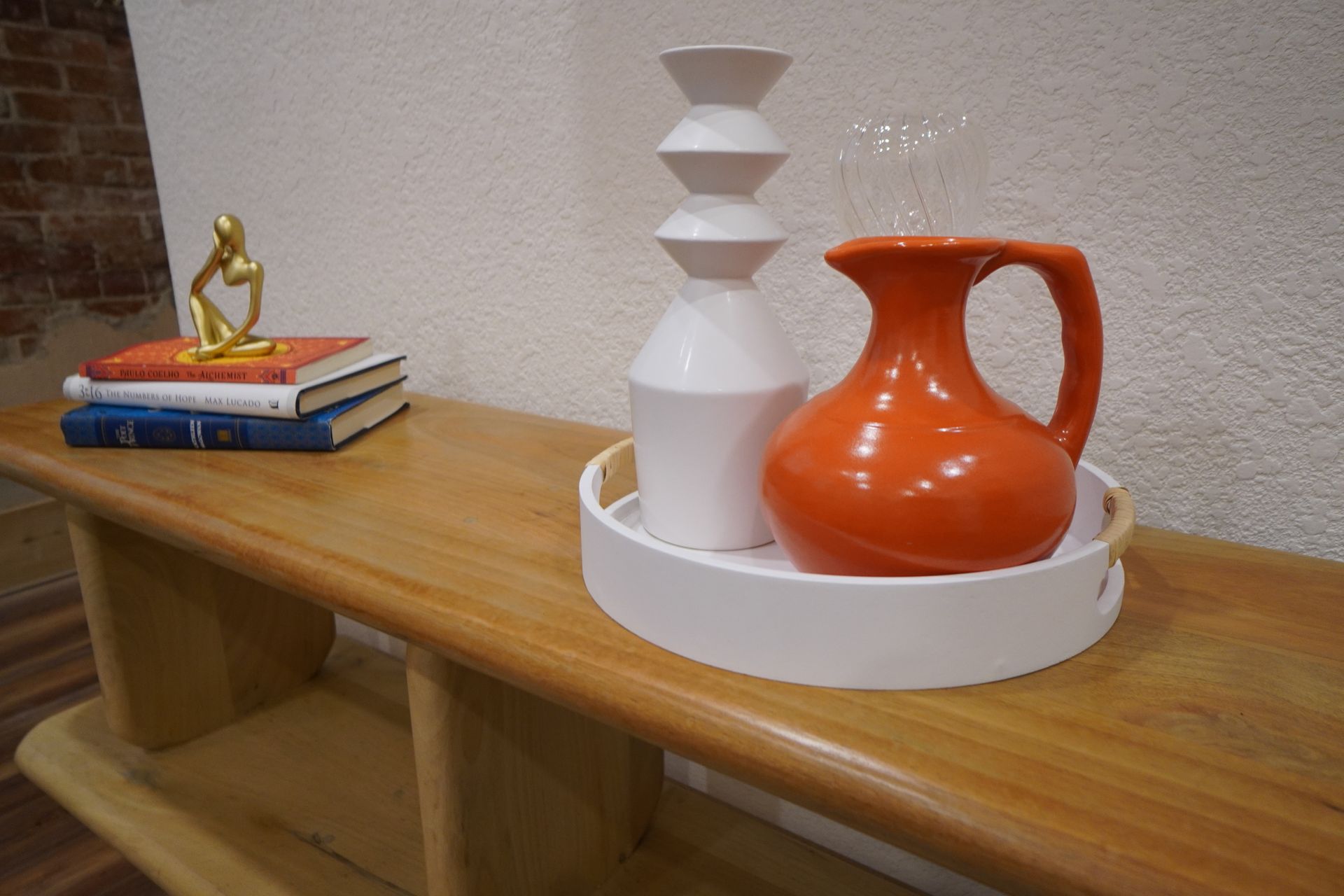 A wooden table holds books, a gold statue, a white tray with a white stacked vase, and an orange pitcher.