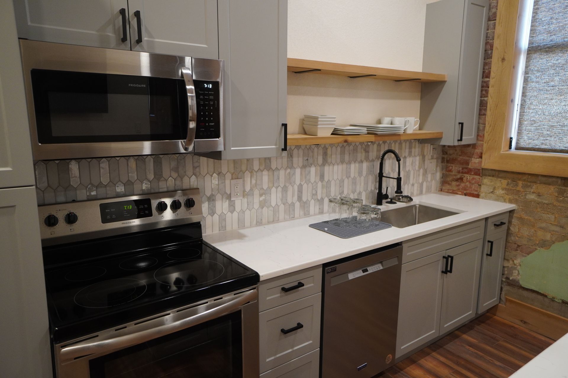Gray and stainless steel kitchen with appliances, backsplash, and countertop, with wood shelving and exposed brick.