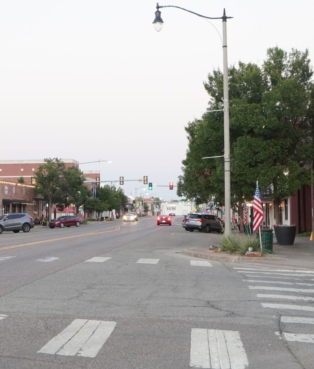 Street scene with cars, crosswalk, trees, buildings, and a streetlight; American flag on the right.