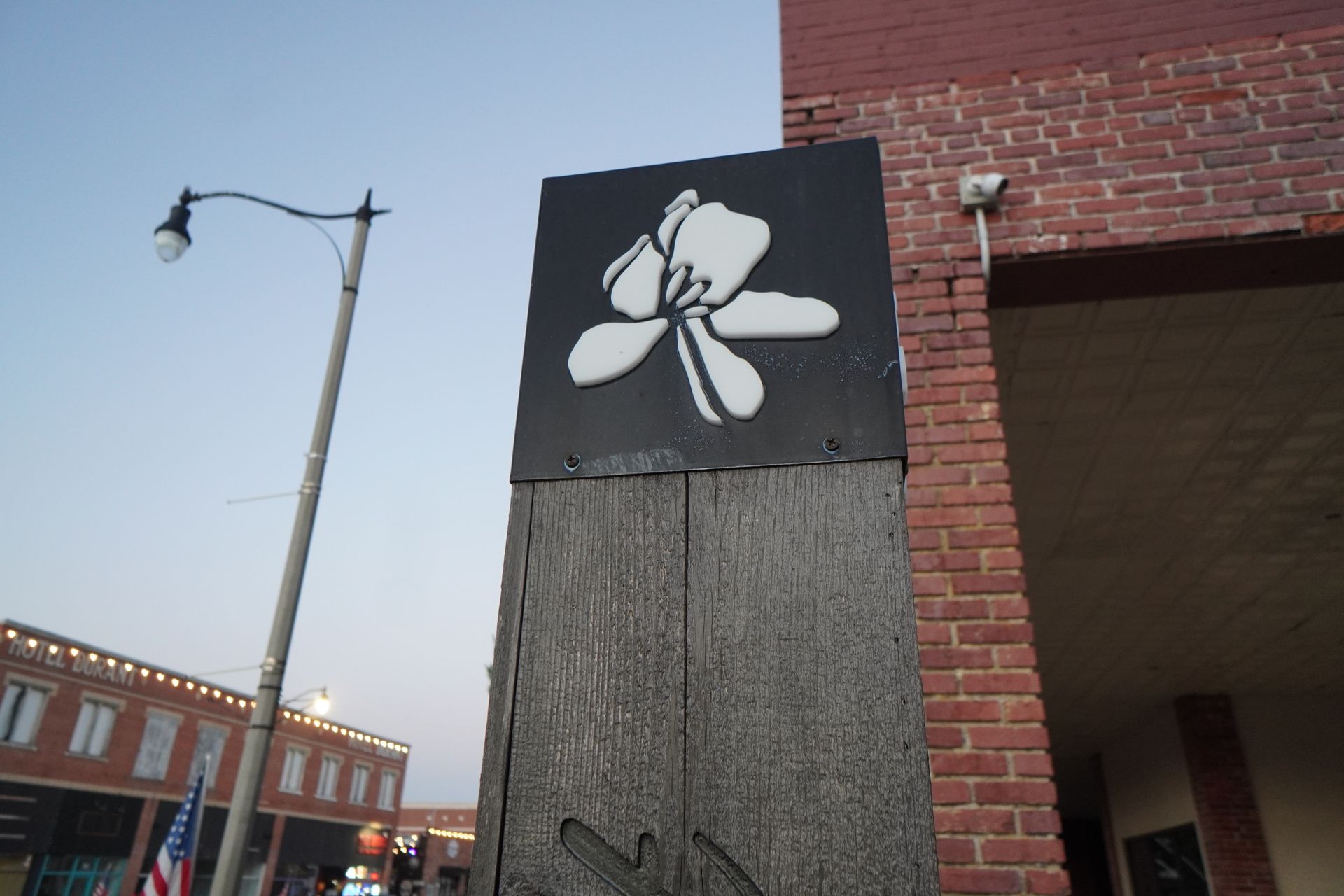 Sign with a white flower on a black square, mounted on a wooden post, in front of a brick building.