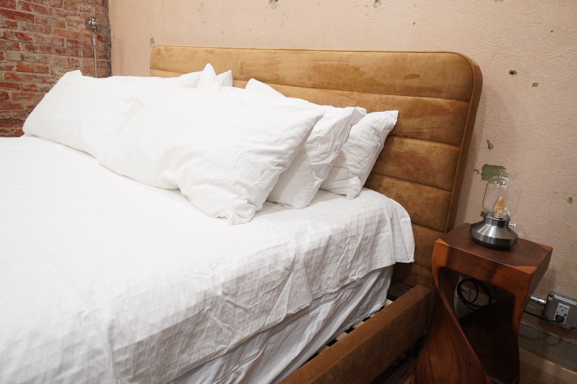 Bed with white bedding and tan headboard, lamp on wooden side table, brick wall in background.
