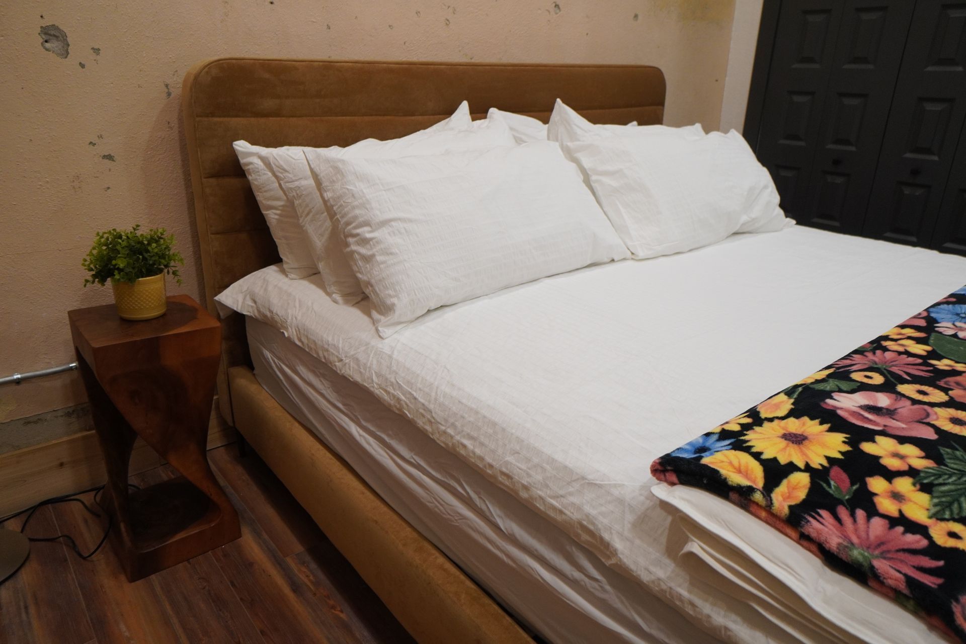 A bed with white bedding, a brown headboard, and a floral blanket. A small wooden table with a plant sits next to the bed.