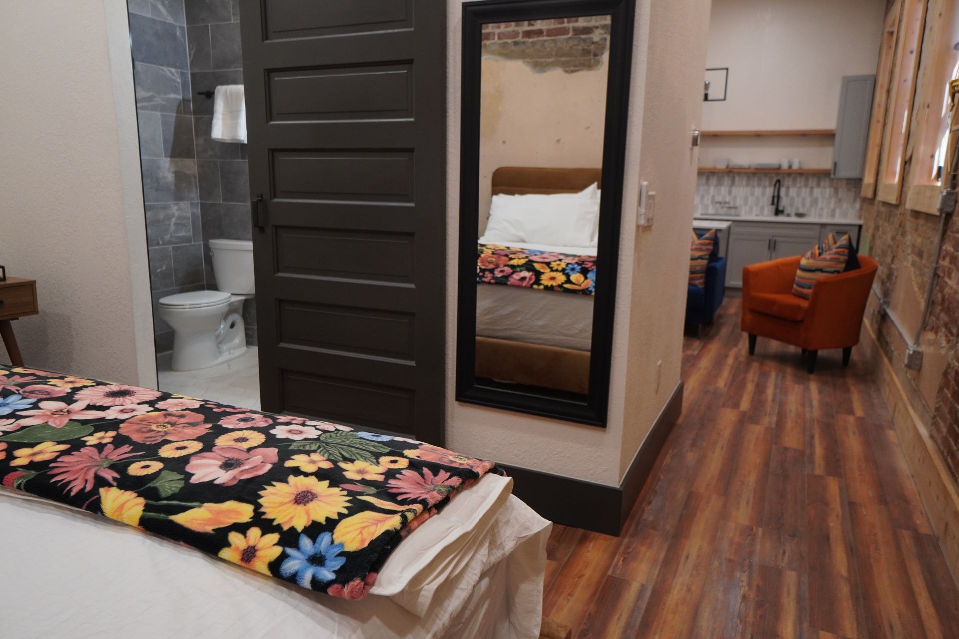 Bedroom with bed, floral blanket, open door to bathroom, mirror, and sitting area with orange chair.