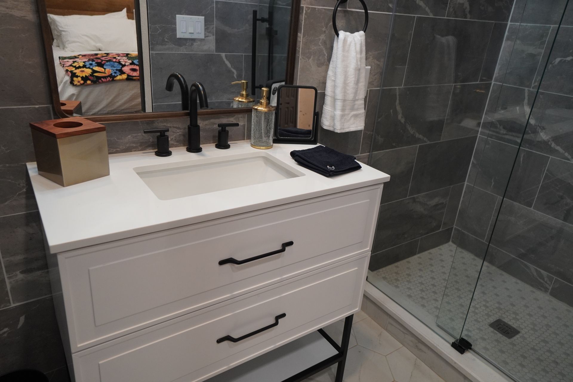 Bathroom vanity with white cabinet, black fixtures, and gray tile. Towel on a ring, and a shower.