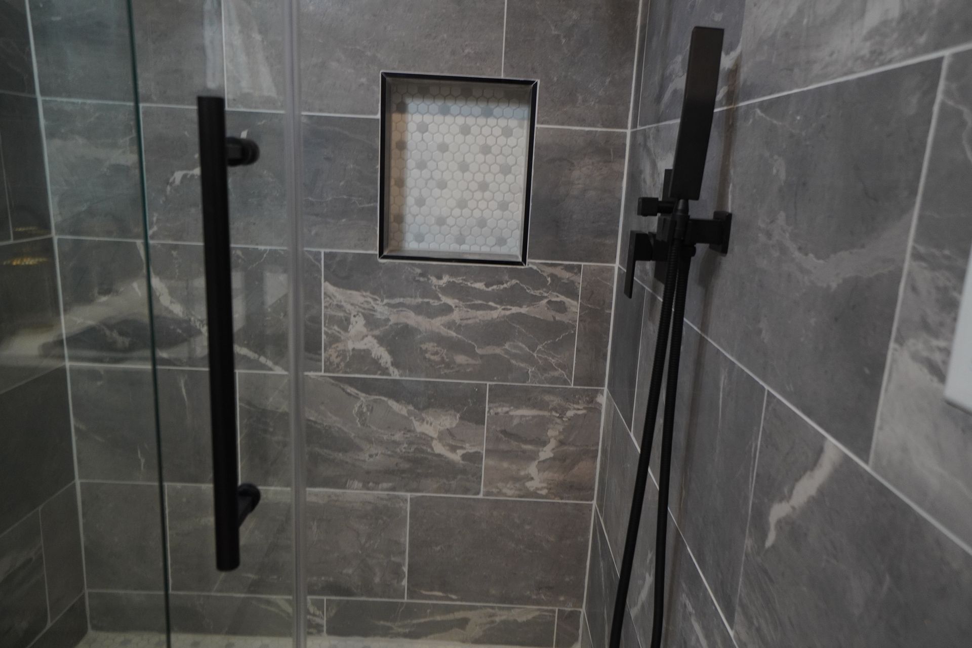 Dark gray tiled shower with black hardware, glass door, and recessed shelf.