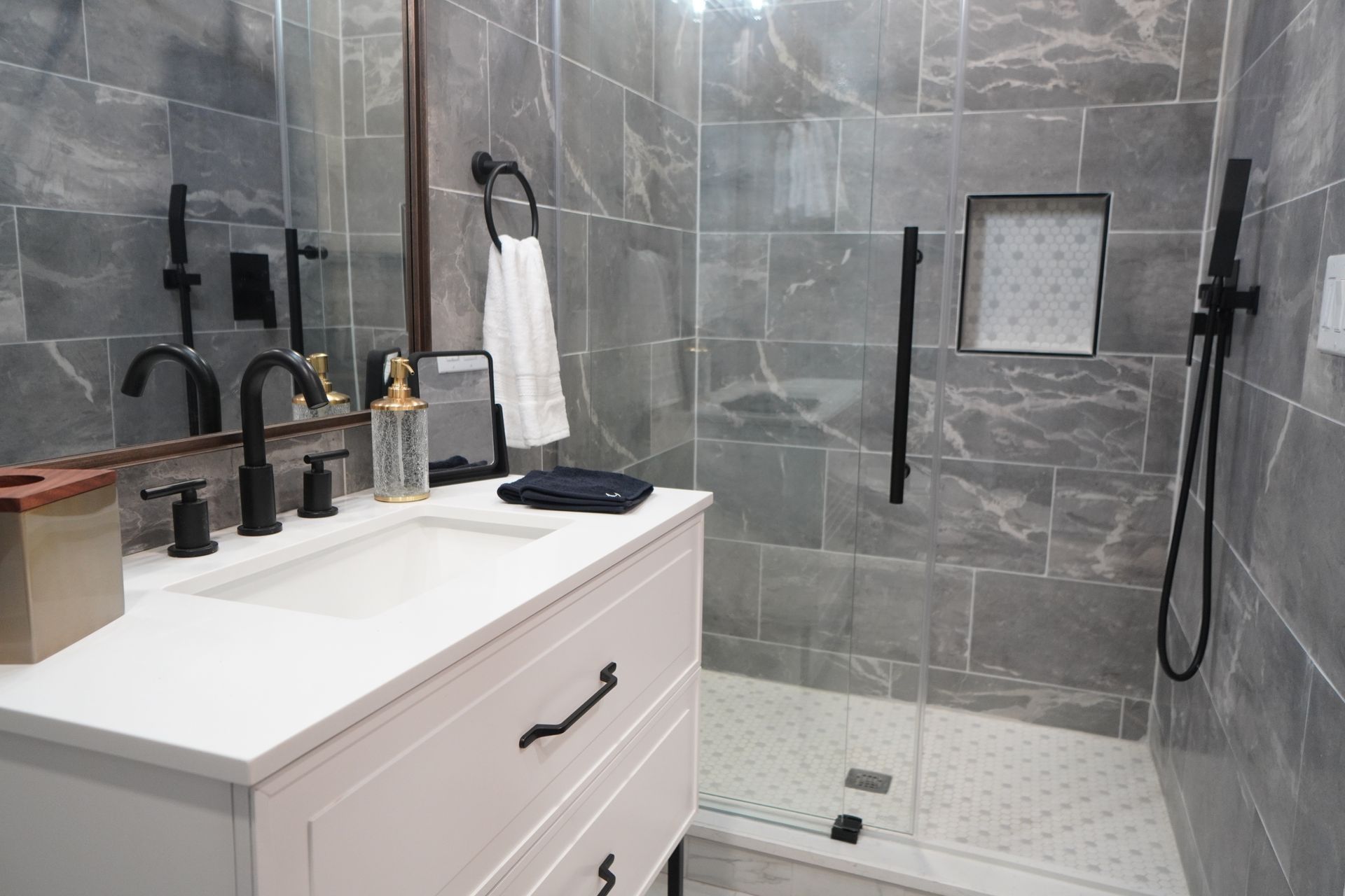 Bathroom with gray tile, white vanity, black fixtures, and glass shower door.