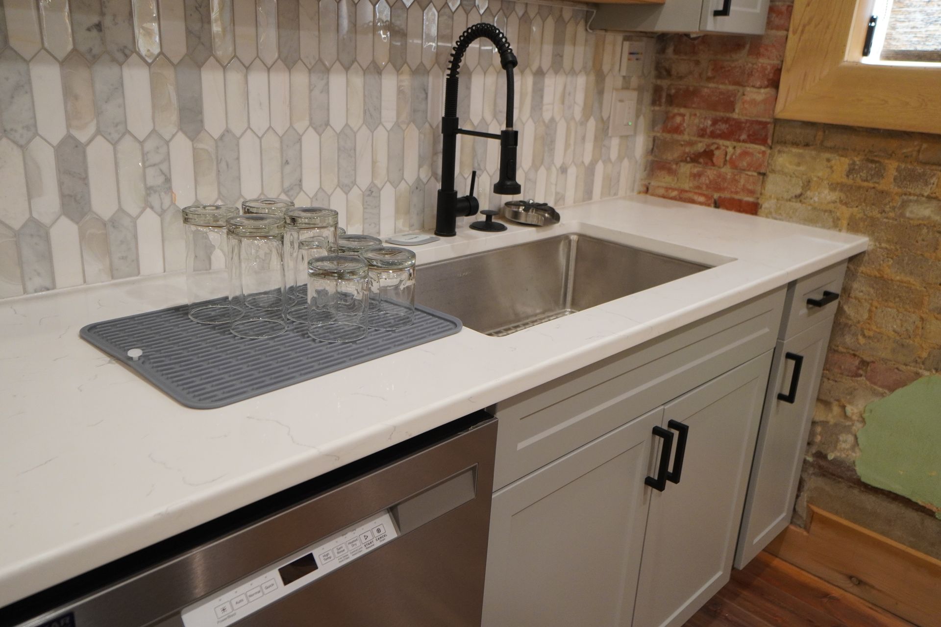 Kitchen sink with black faucet, light cabinets, and a white countertop. Glasses on a drying mat.
