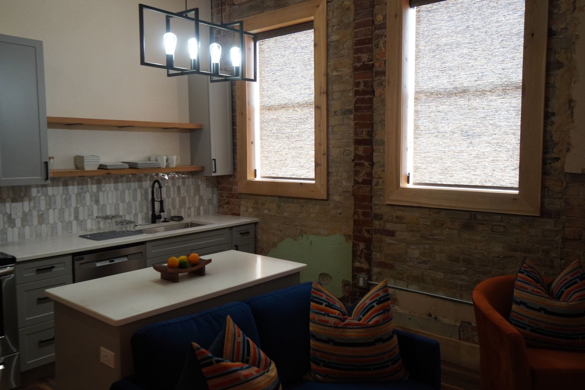 Kitchen area with island, windows, and brick walls. Modern pendant light hangs above the sink.