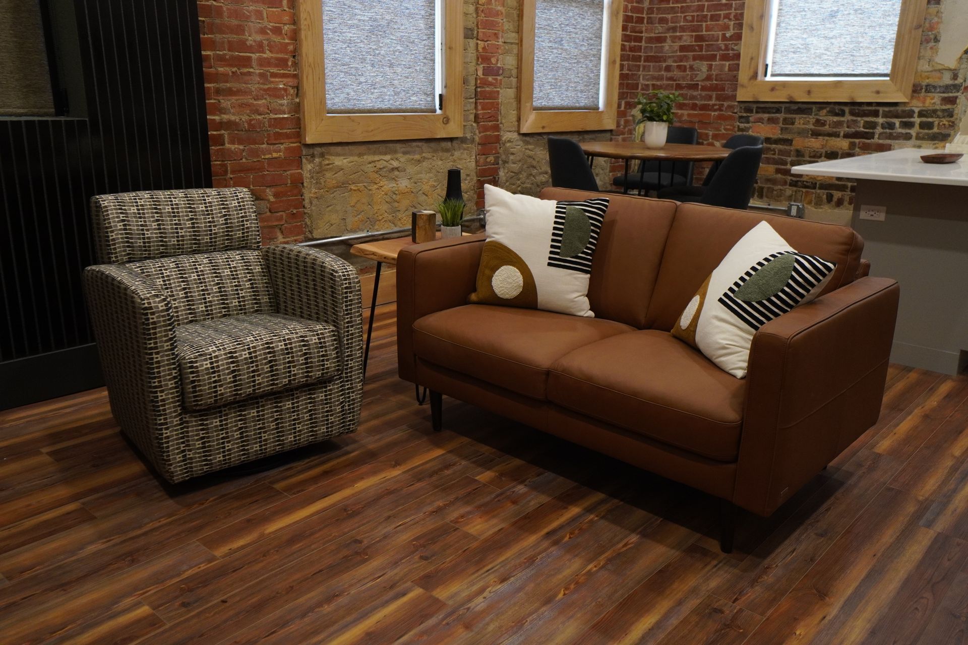 Living room with brown leather sofa, patterned armchair, exposed brick walls, and hardwood floor.