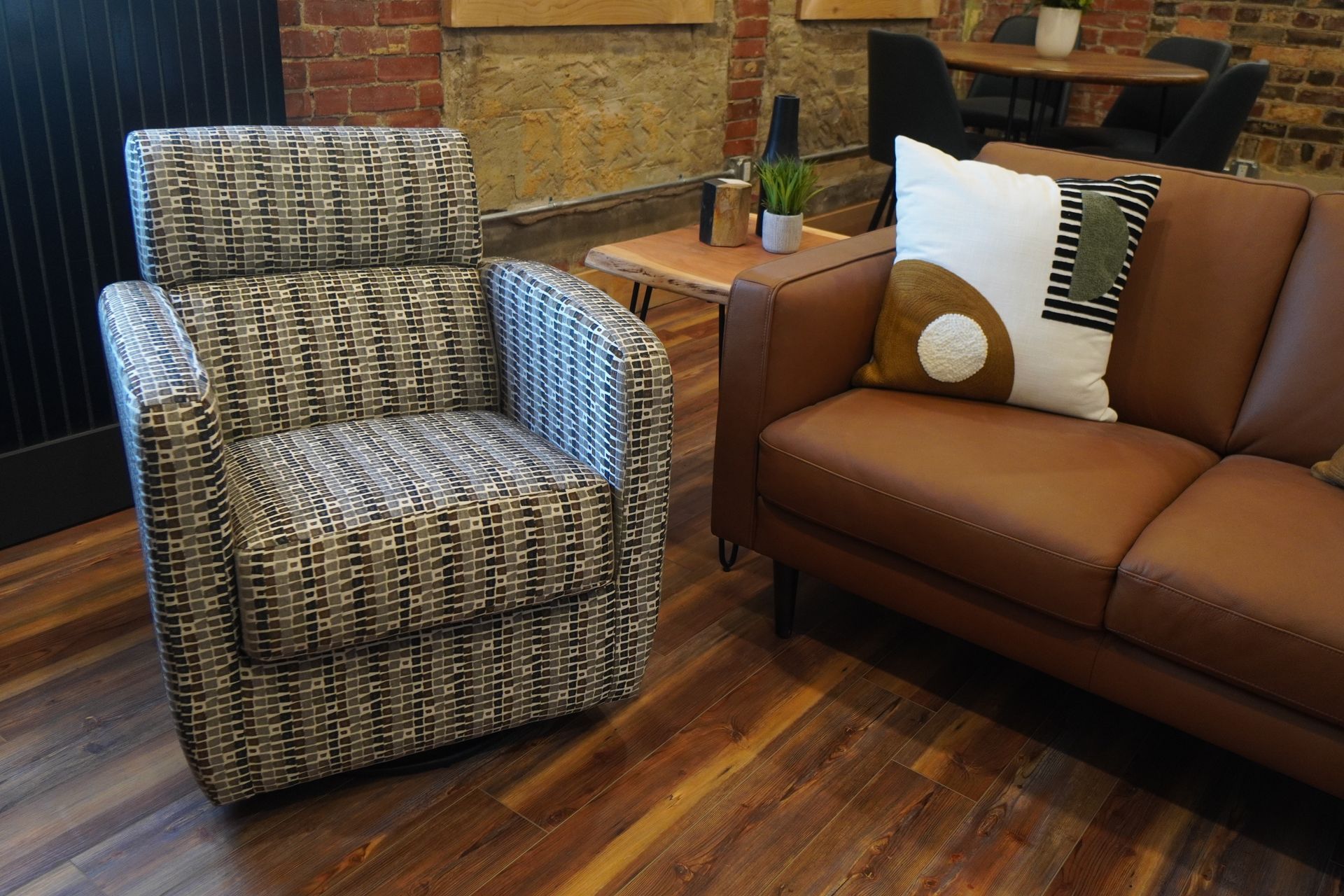 Armchair with patterned fabric beside a brown leather sofa. Coffee table with plant and brick wall background.