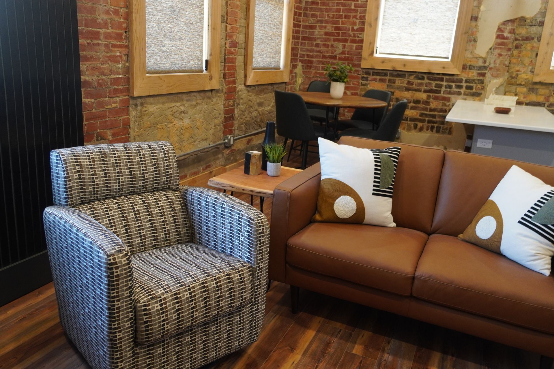 Cozy living space with brown leather sofa, patterned armchair, and exposed brick walls. Dining table in the background.