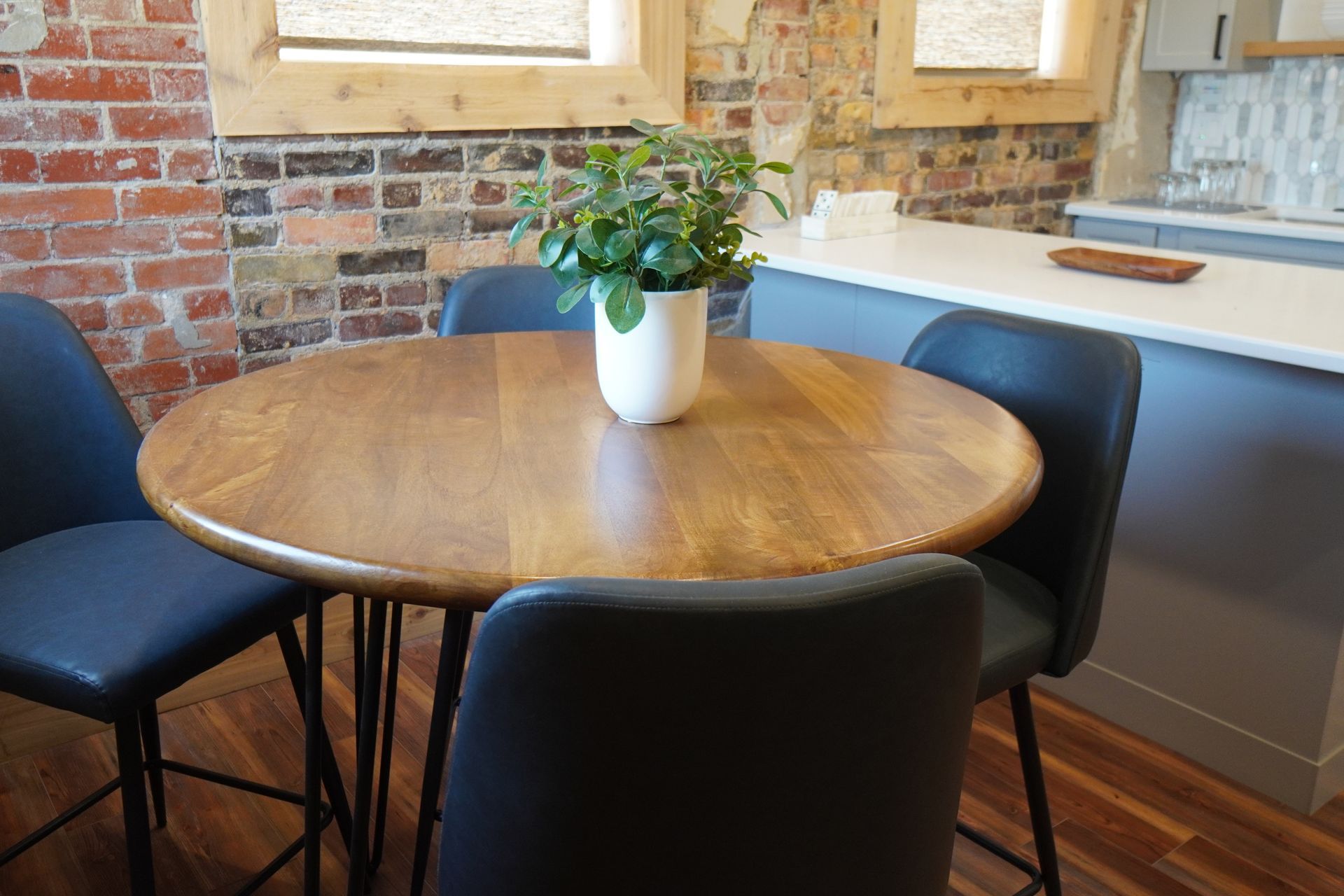 Round wooden table with four black chairs and a plant in a white pot in front of a brick wall and kitchen.