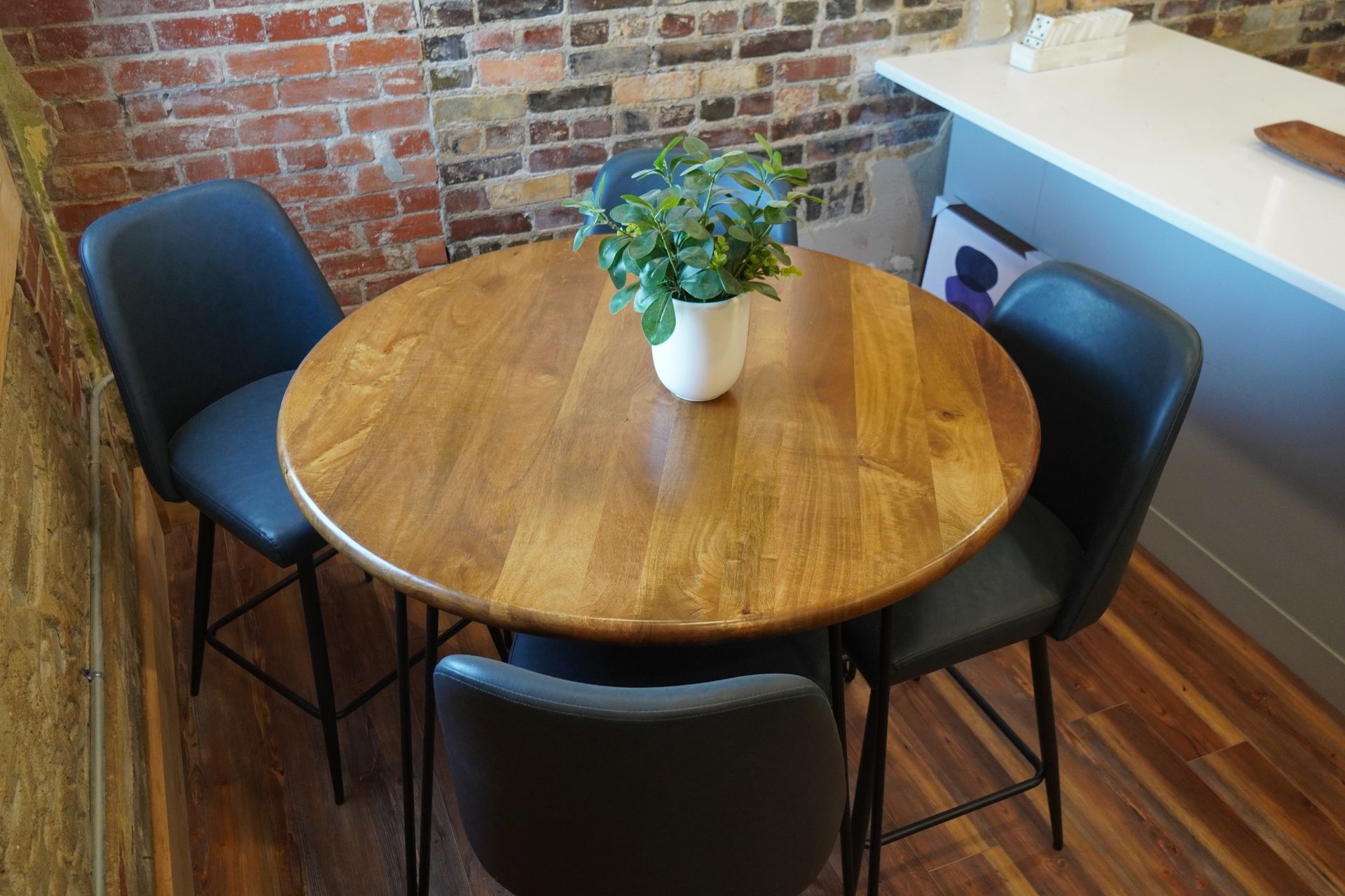 Round wooden table with four black chairs in front of a brick wall, topped with a plant.