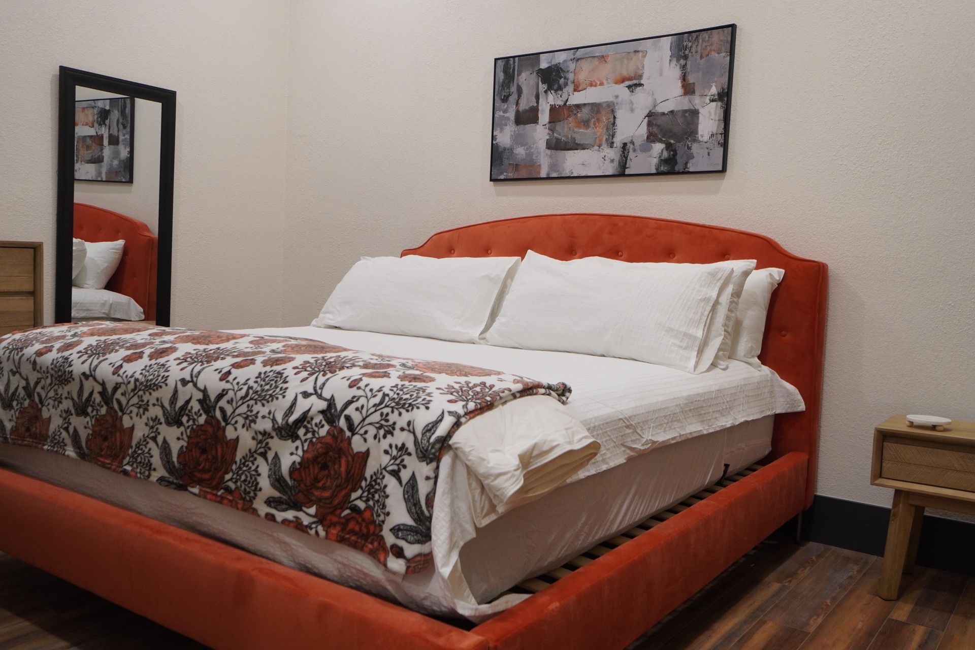 An orange bed with white bedding, floral blanket, mirror, artwork, and a nightstand in a bedroom.