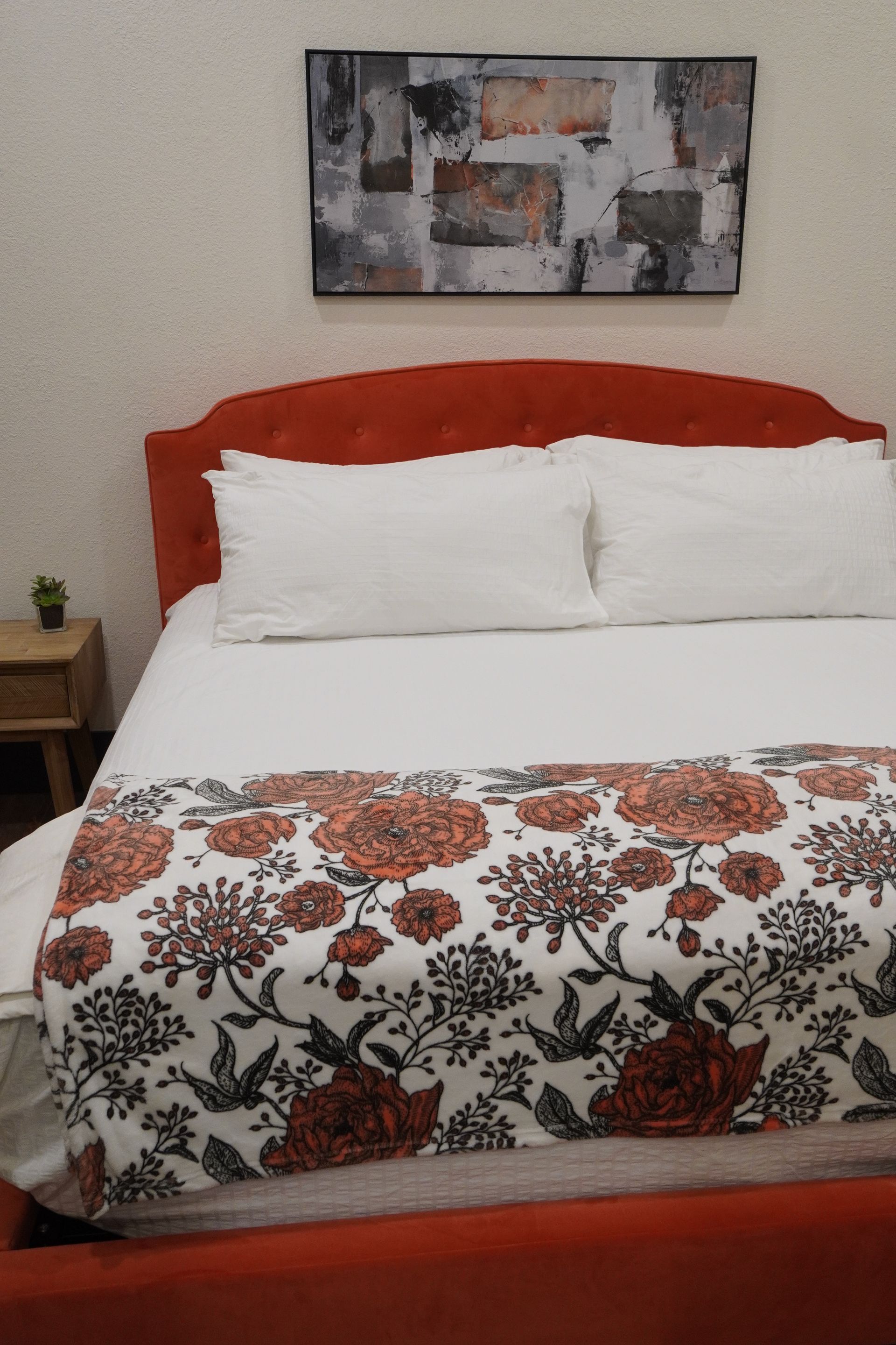 Orange bed with white bedding, floral runner, and abstract painting above. A small nightstand is on the left.