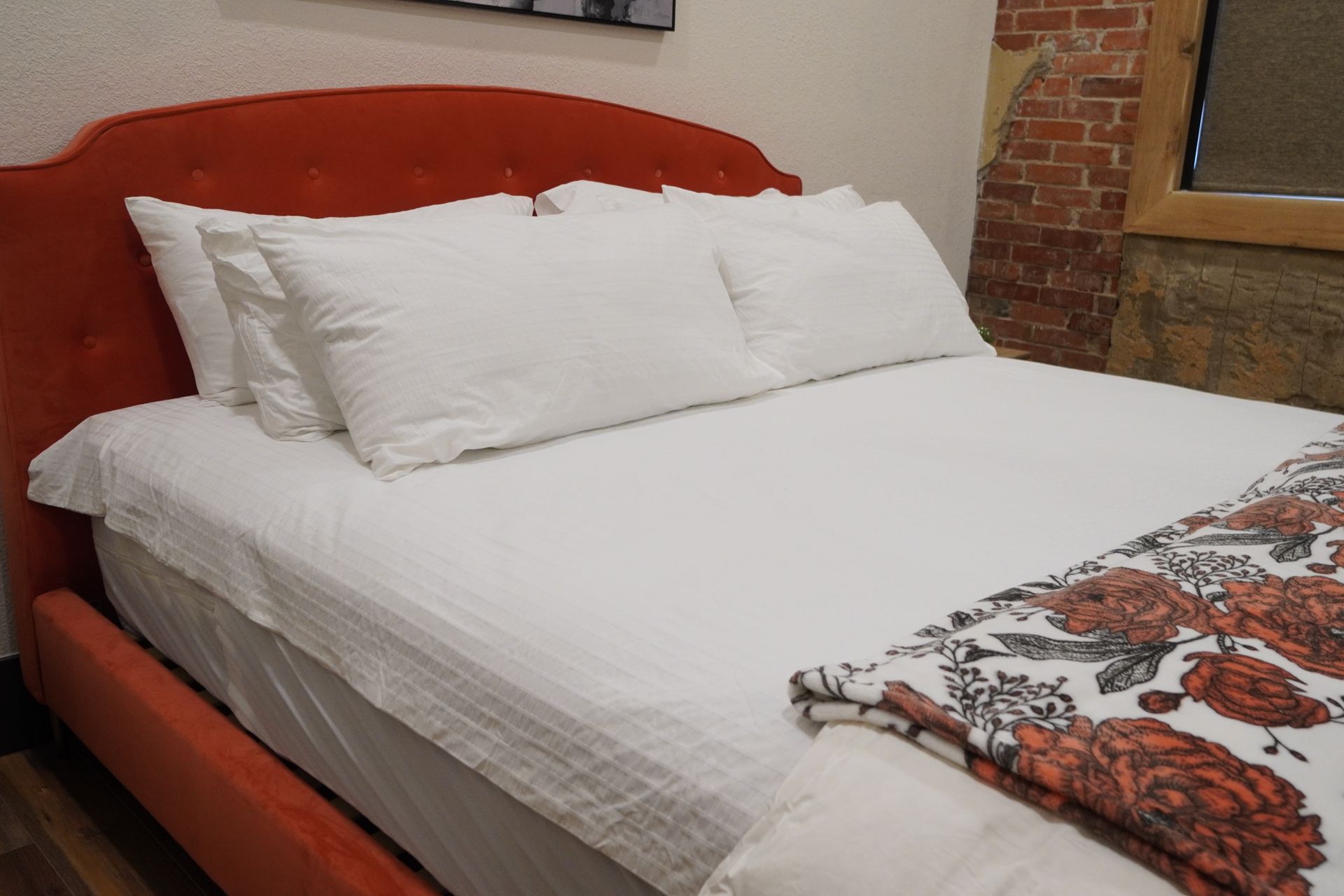 Red velvet bed with white linens and floral throw, against a brick wall.
