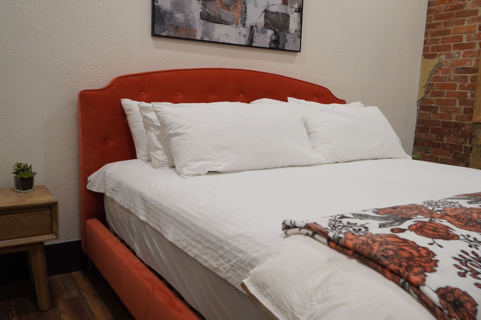 A bed with an orange headboard, white bedding, and floral blanket. A nightstand and brick wall are visible.