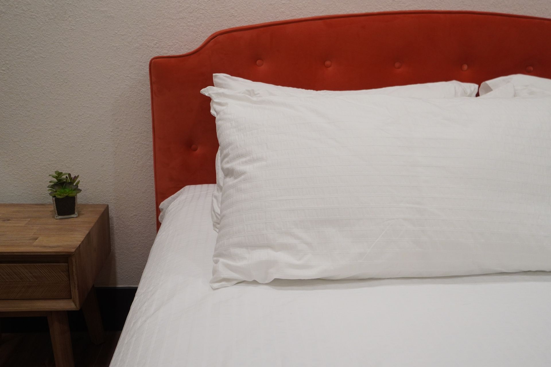 Bed with white sheets, large pillow, orange headboard, and wooden nightstand with plant.