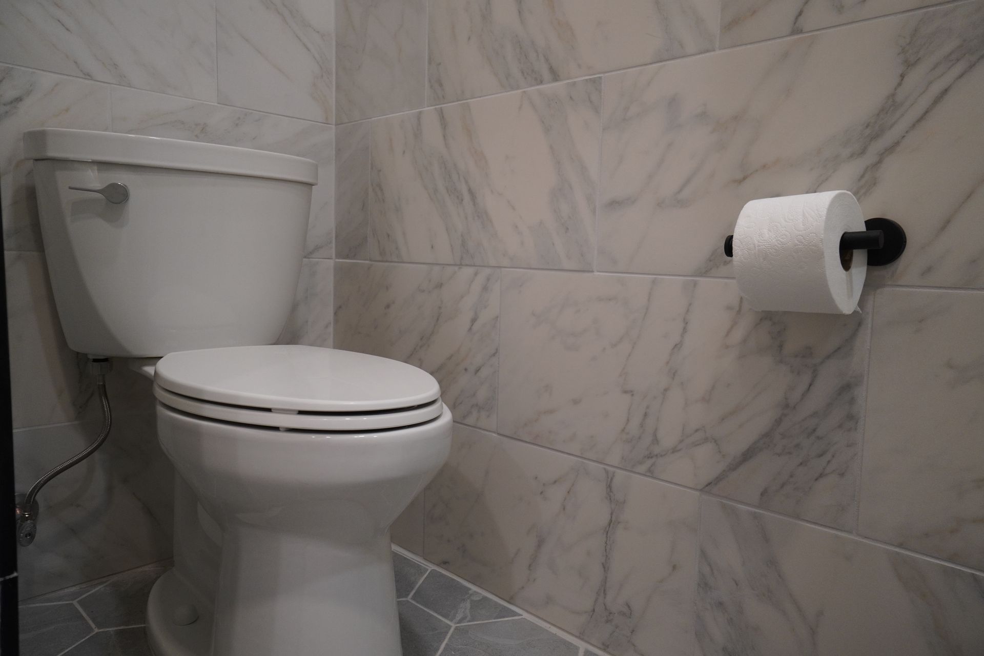 White toilet next to a wall-mounted toilet paper holder. Marble-like wall tiles.