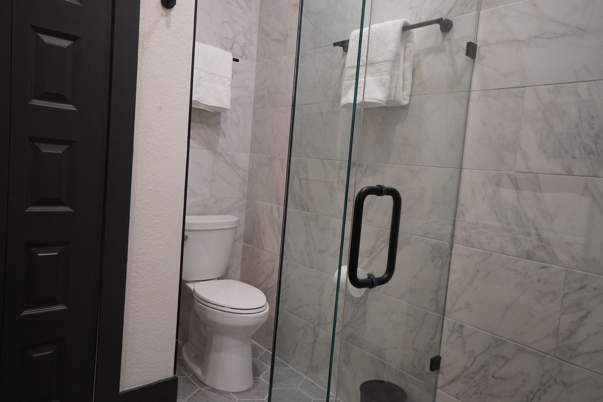 Bathroom with glass shower, toilet, and towel racks. Walls are tiled in gray. Black door on left.