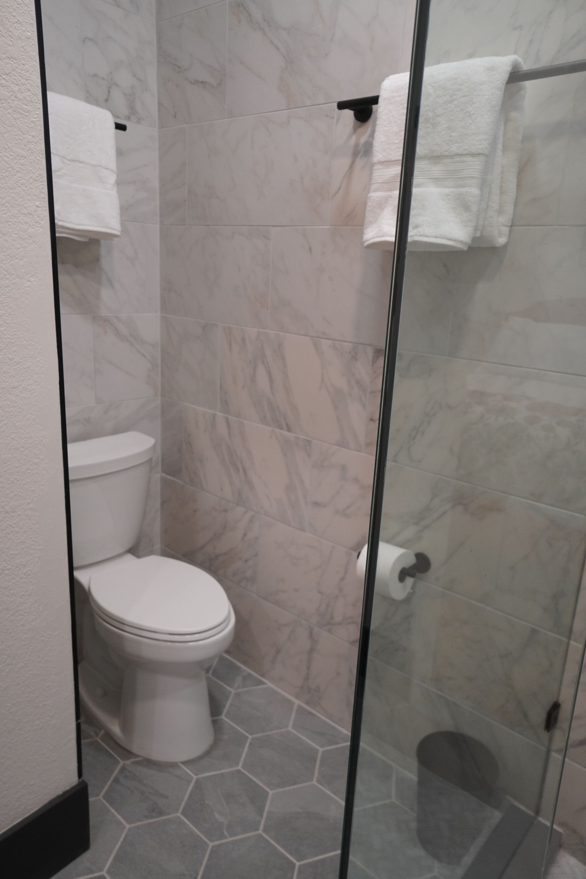 Bathroom with a toilet, gray and white marble-like tile, hexagon floor tiles, glass shower door, and towels on black bars.