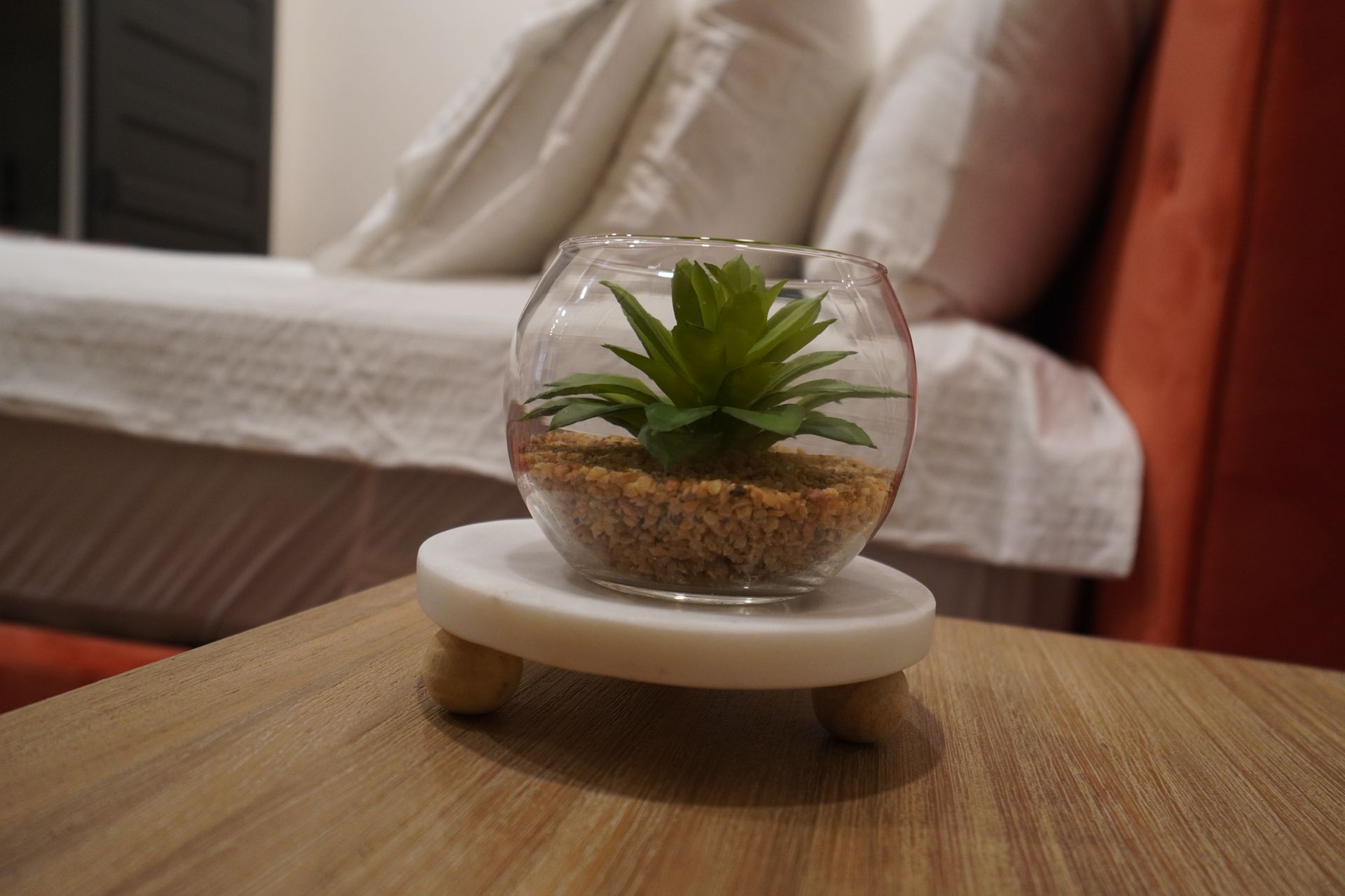 Glass terrarium with a succulent on a wooden stand, on a nightstand beside a bed with white bedding.