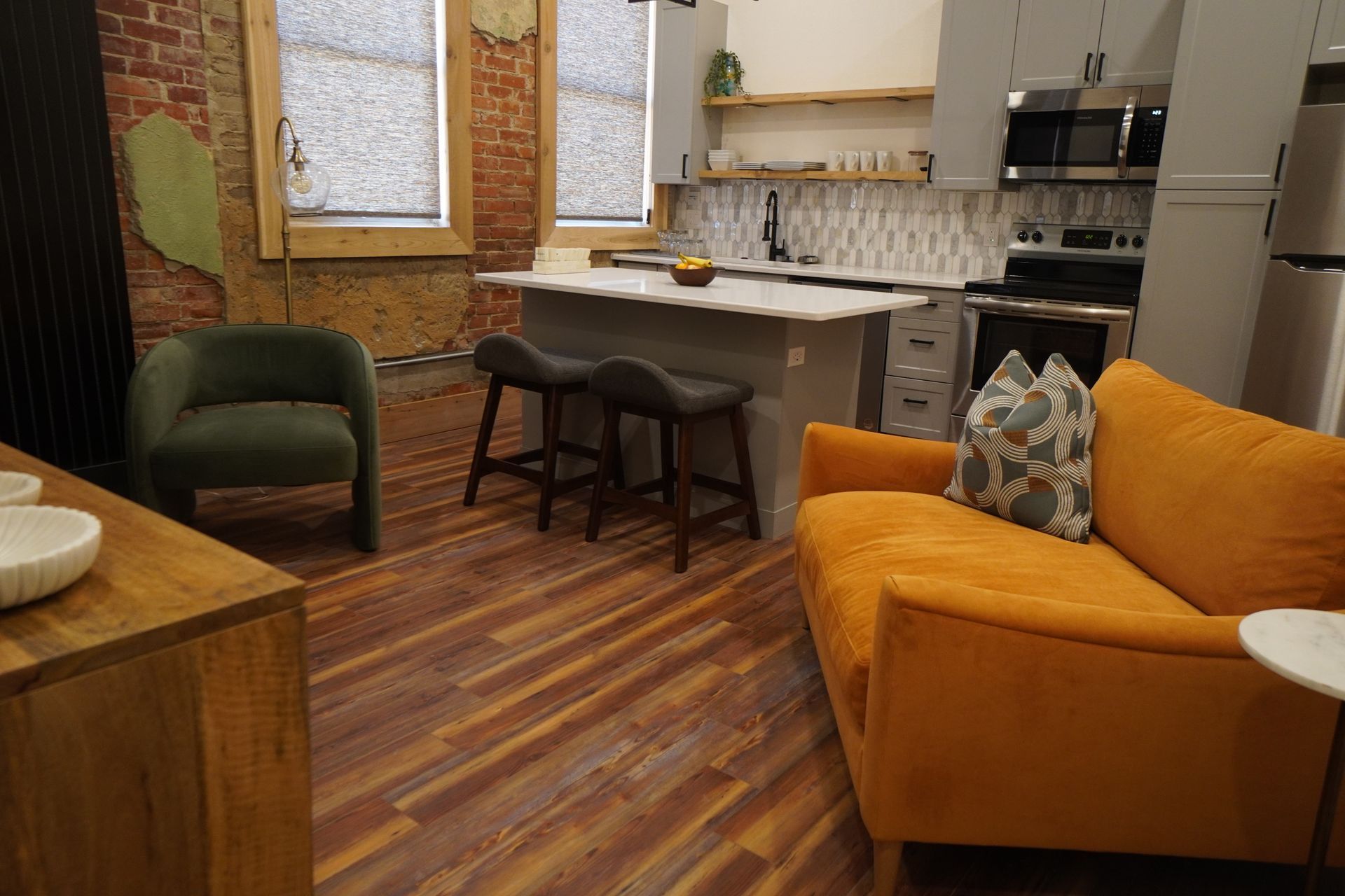 Cozy kitchen and living area with orange couch, green armchair, island with stools, and exposed brick.