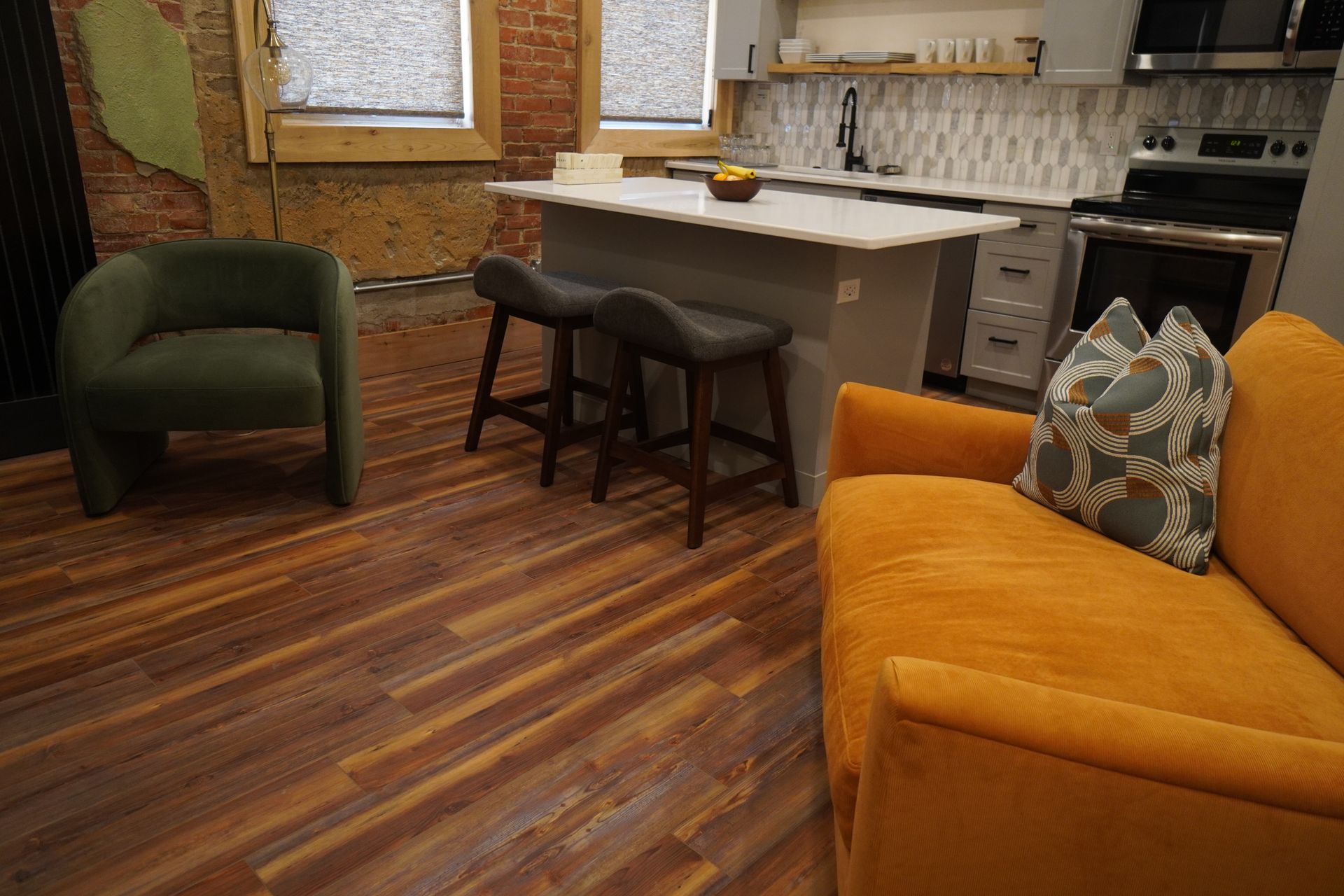 Cozy living area with an orange couch, green chair, and kitchen island with stools. Brick wall and wood floors.