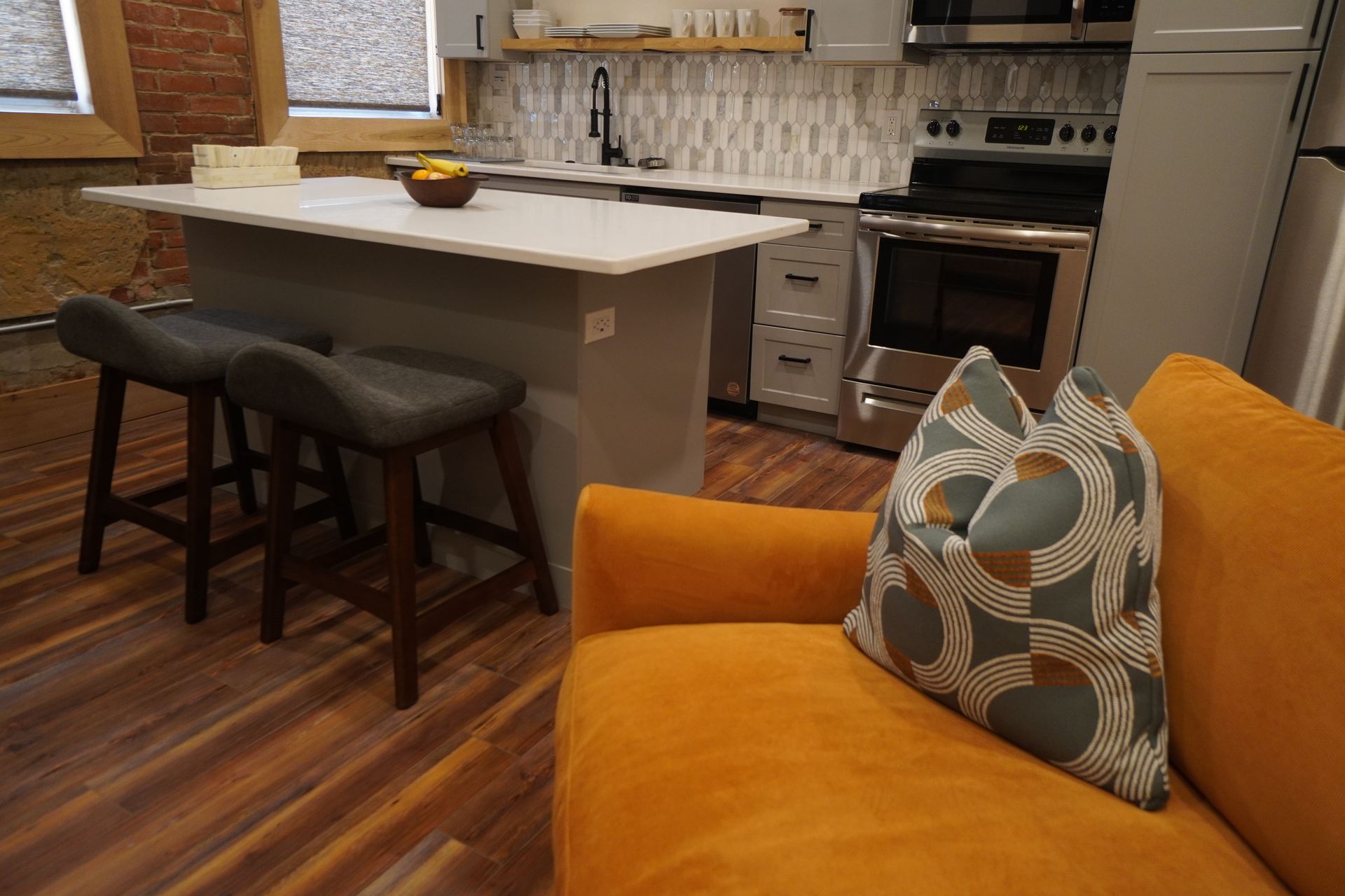 An open kitchen with a sofa. The kitchen has a gray island with bar stools. The sofa is orange with a patterned pillow.