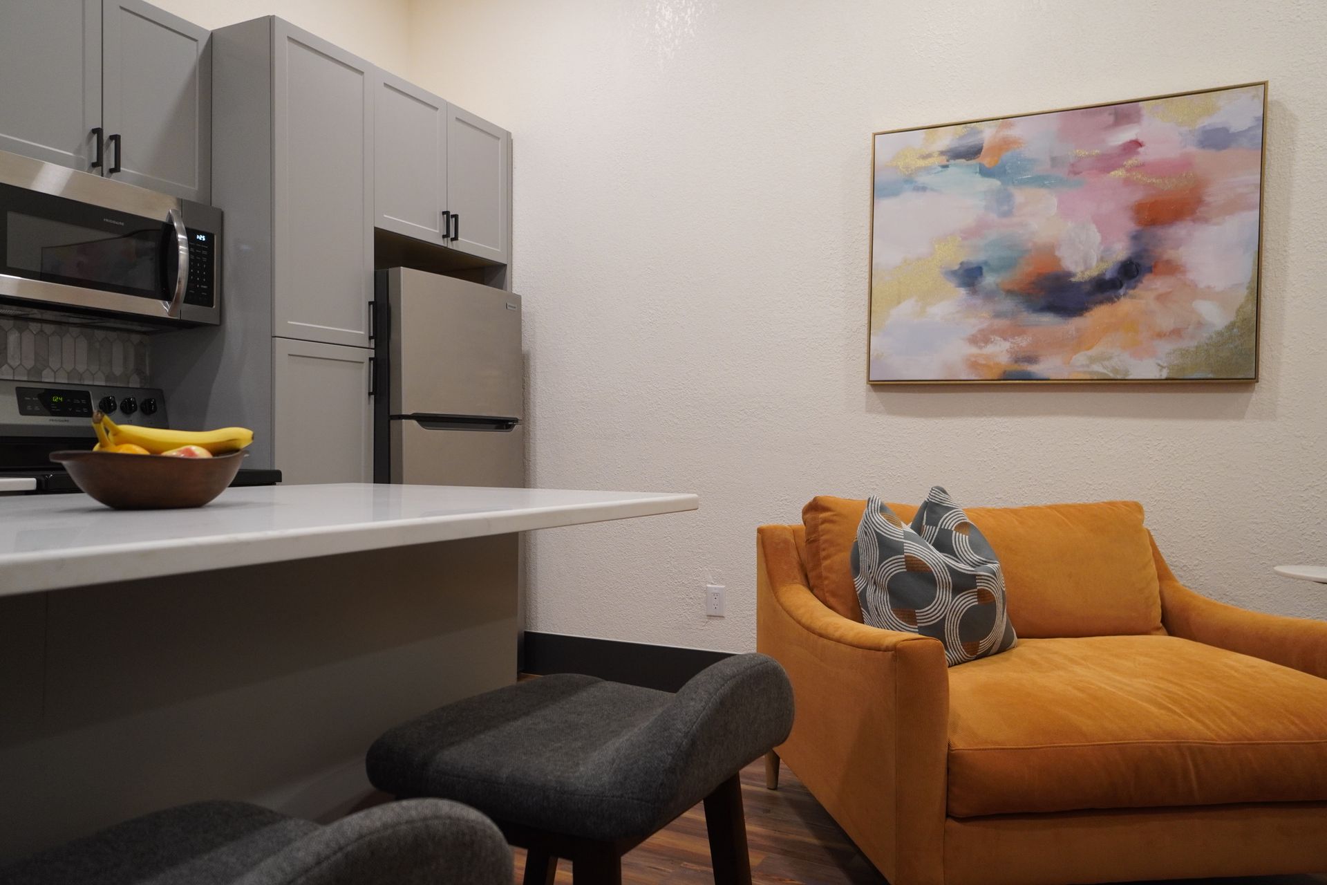 Kitchen and seating area. Gray cabinets, stainless steel appliances, orange chaise lounge, artwork, and bar stools.