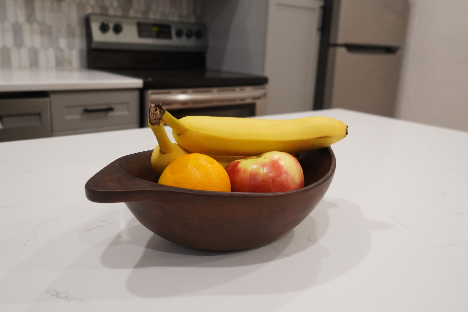 Wooden bowl of fruit (banana, orange, apple) on a white kitchen countertop with stove and refrigerator in background.
