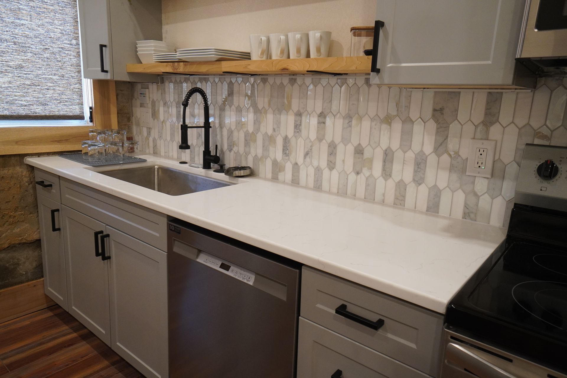 Gray and white kitchen with a stainless steel sink, dishwasher, and stove; a wooden shelf holds dishes.