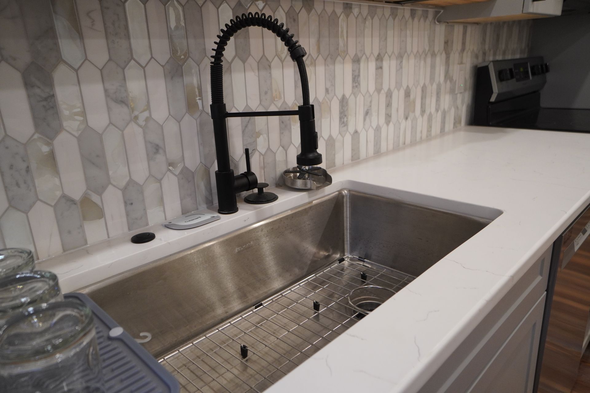 Stainless steel kitchen sink with black faucet, white countertop, and patterned backsplash.