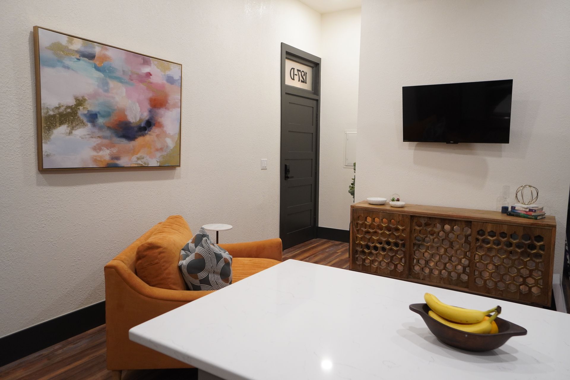 Living room with orange chair, white countertop, artwork, TV, and dark door labeled 