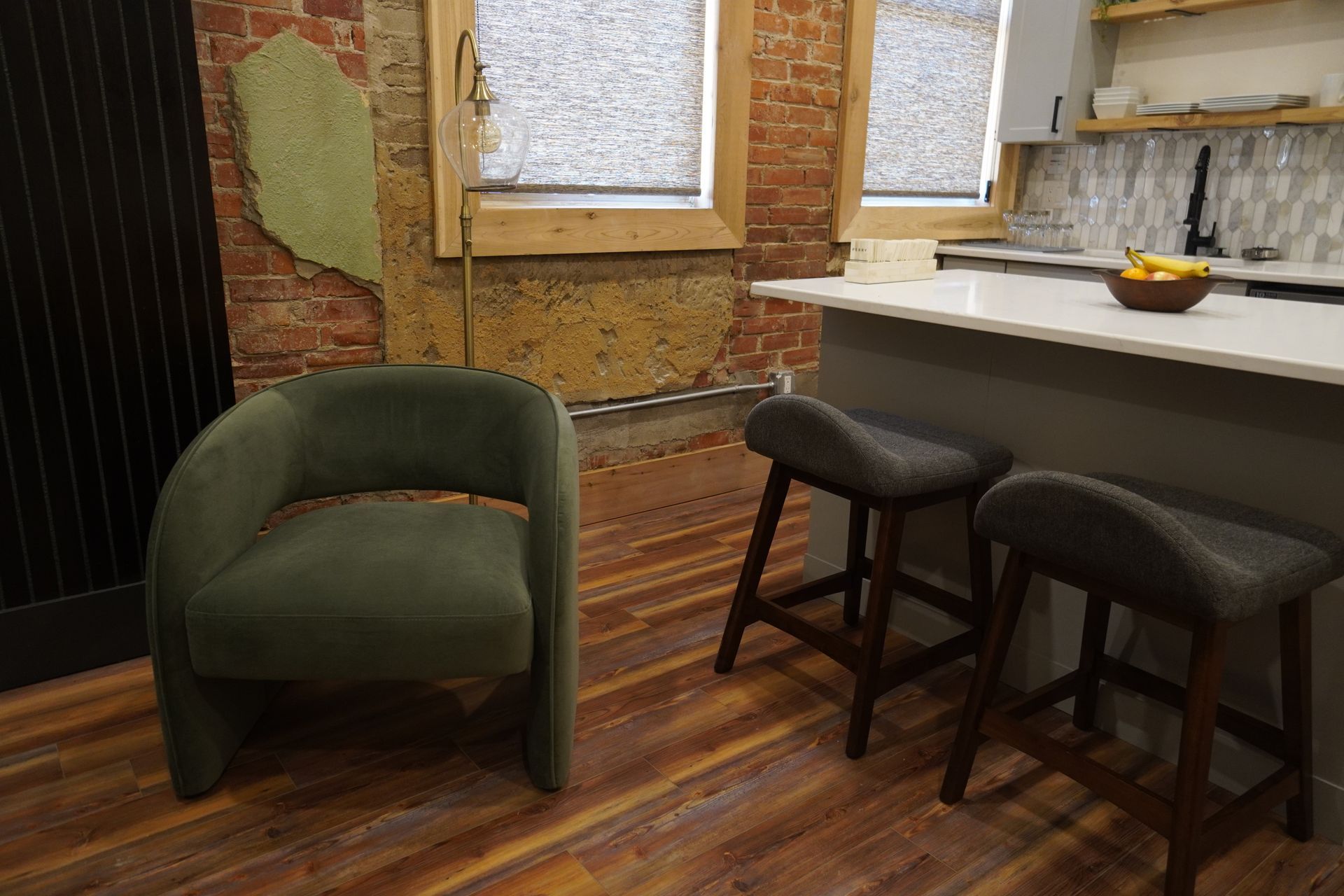 Green armchair beside two bar stools in a kitchen with exposed brick and a small island.