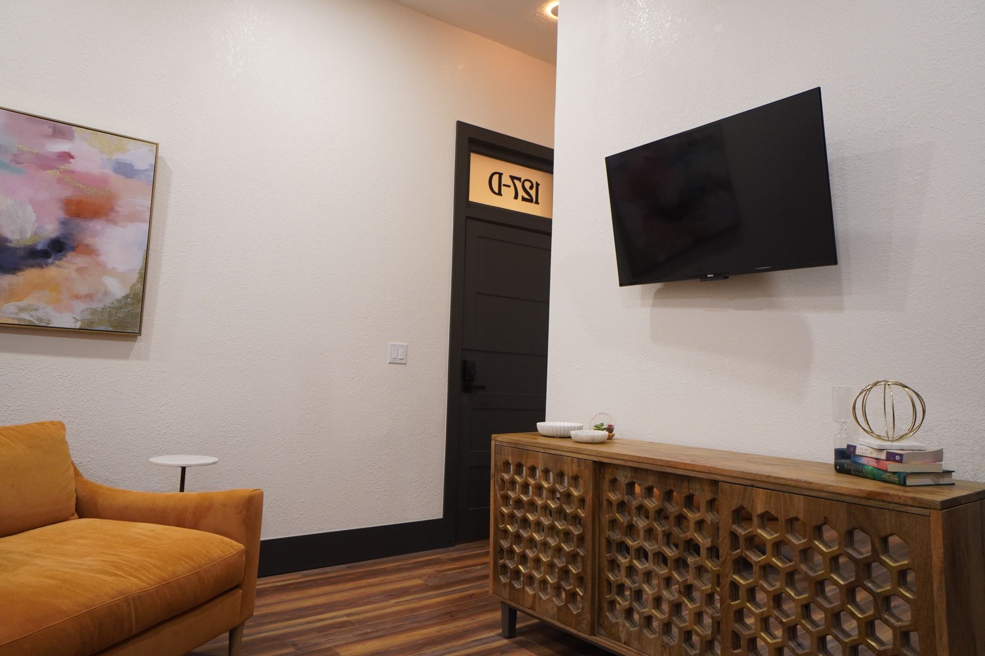 Living room with orange couch, wooden cabinet, and mounted TV. Door with room number.