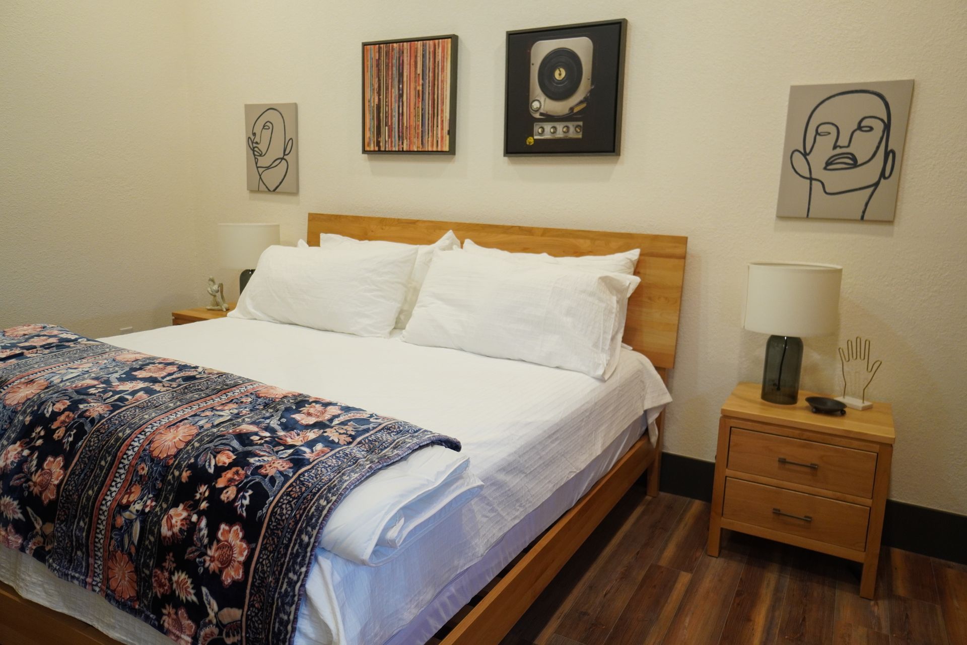 Bedroom with wooden bed frame, white bedding, floral blanket, nightstands, and art on the wall.