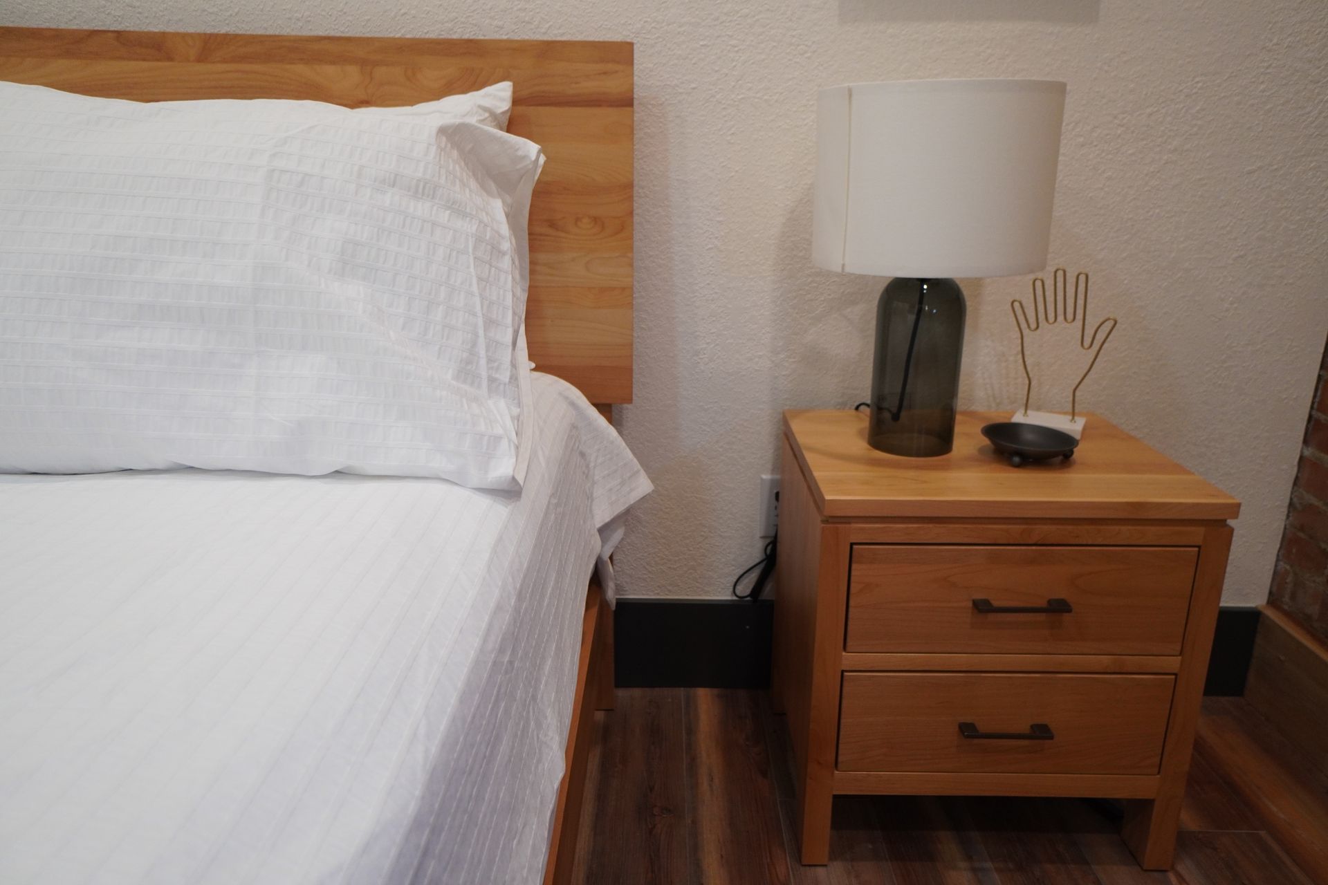 Bed with white bedding next to a wooden nightstand with a lamp and decor.