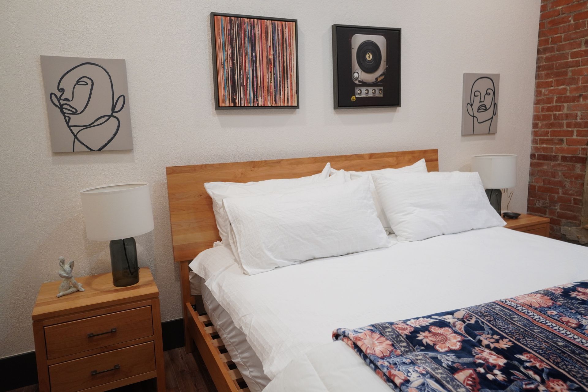 Bedroom with bed, nightstands, artwork, and brick wall. White bedding, wooden furniture, and lamps.