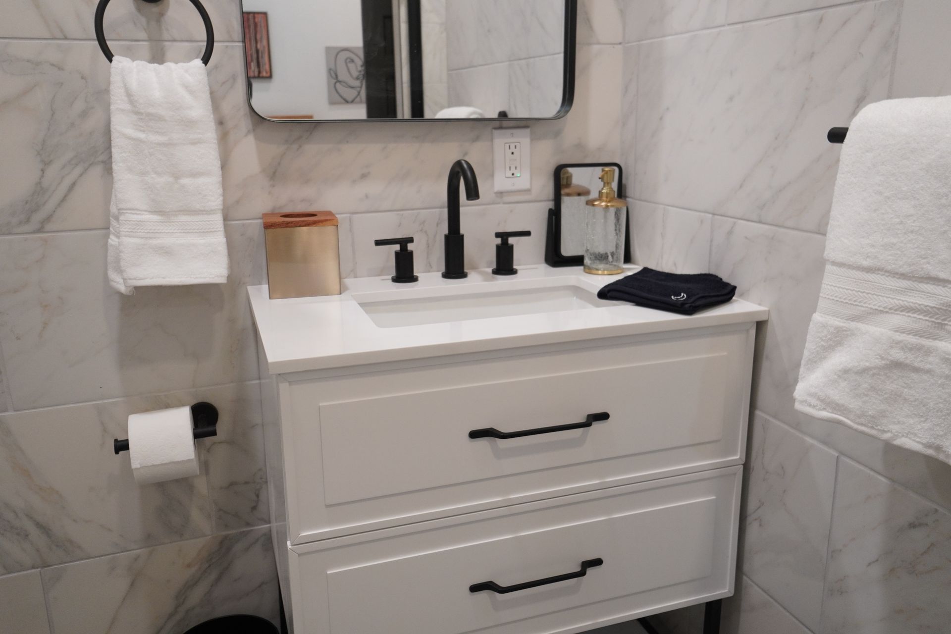White bathroom vanity with black hardware, towels, and mirror on marble tile.