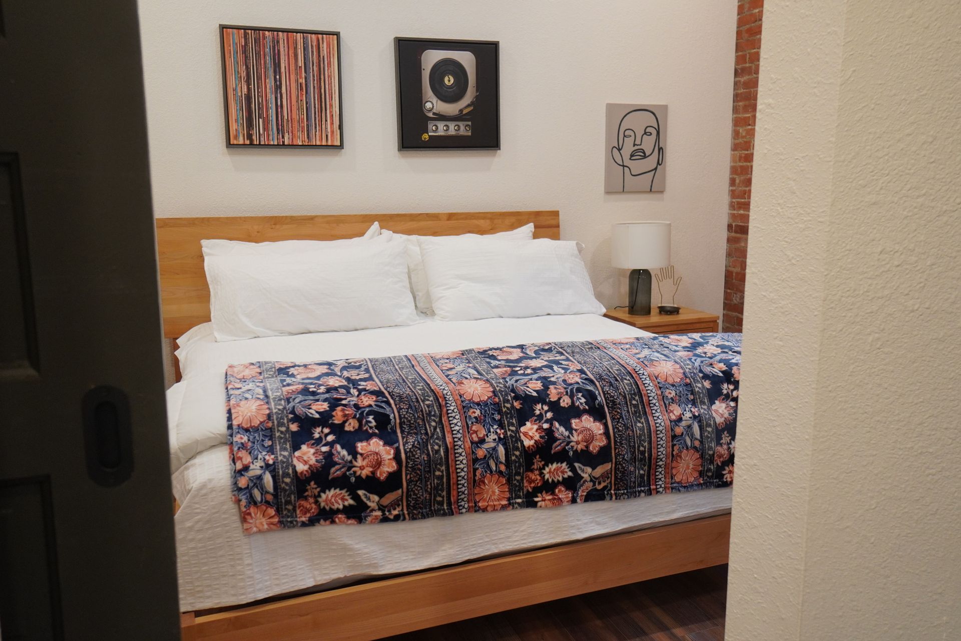Bedroom with wooden bed, white bedding, floral blanket, art on walls, and brick column.