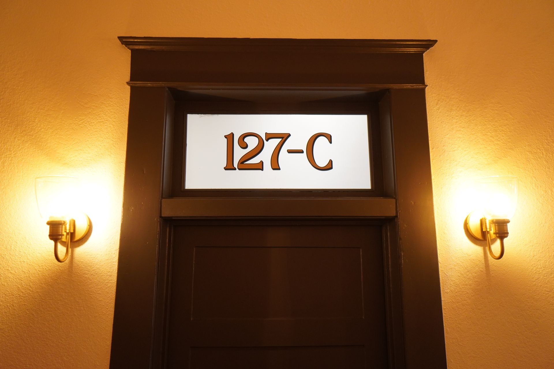 Doorway with number 127-C above, flanked by sconces on a warm-toned wall.