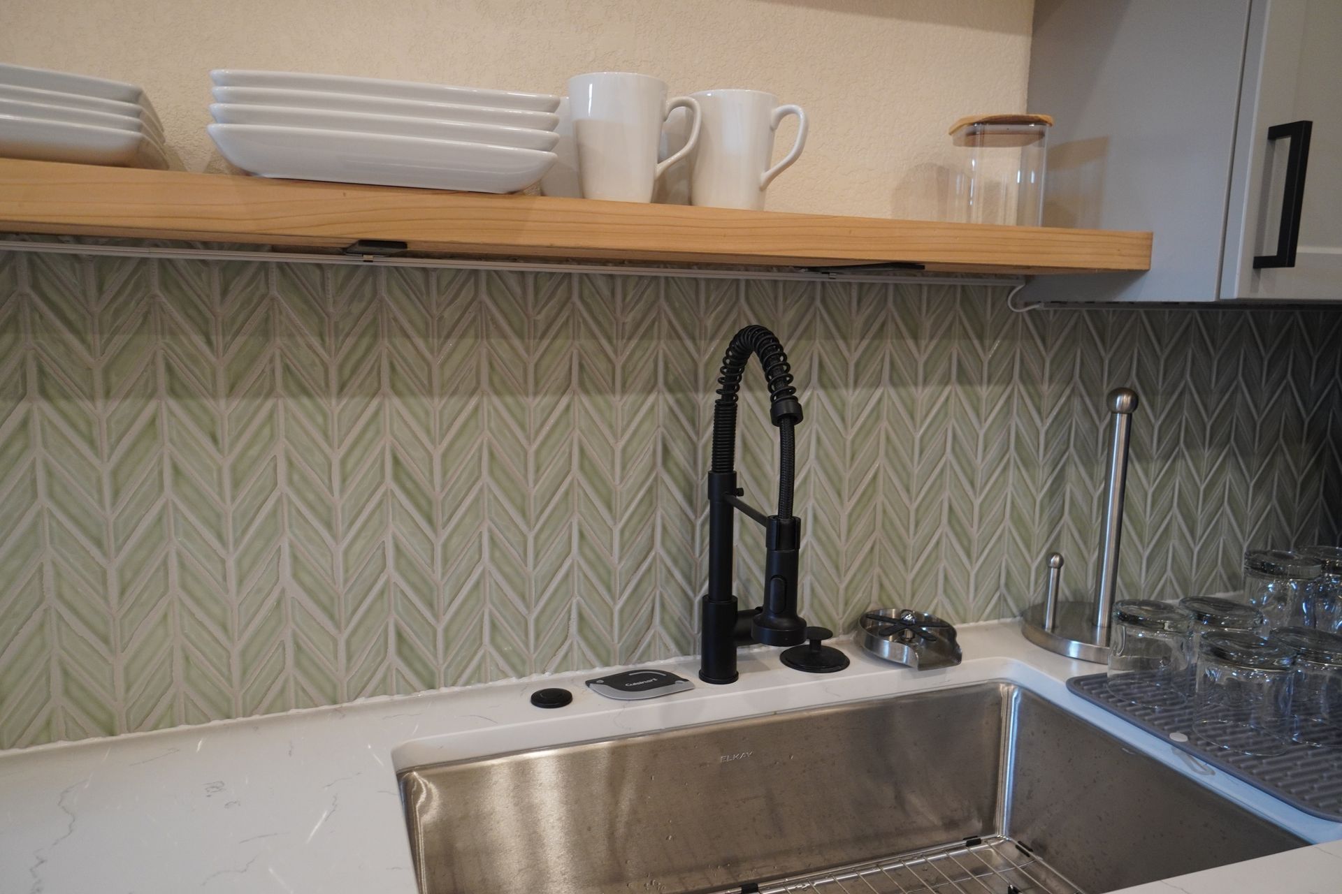 Kitchen with stainless steel sink, black faucet, and patterned tile backsplash. Shelves hold dishes and mugs.