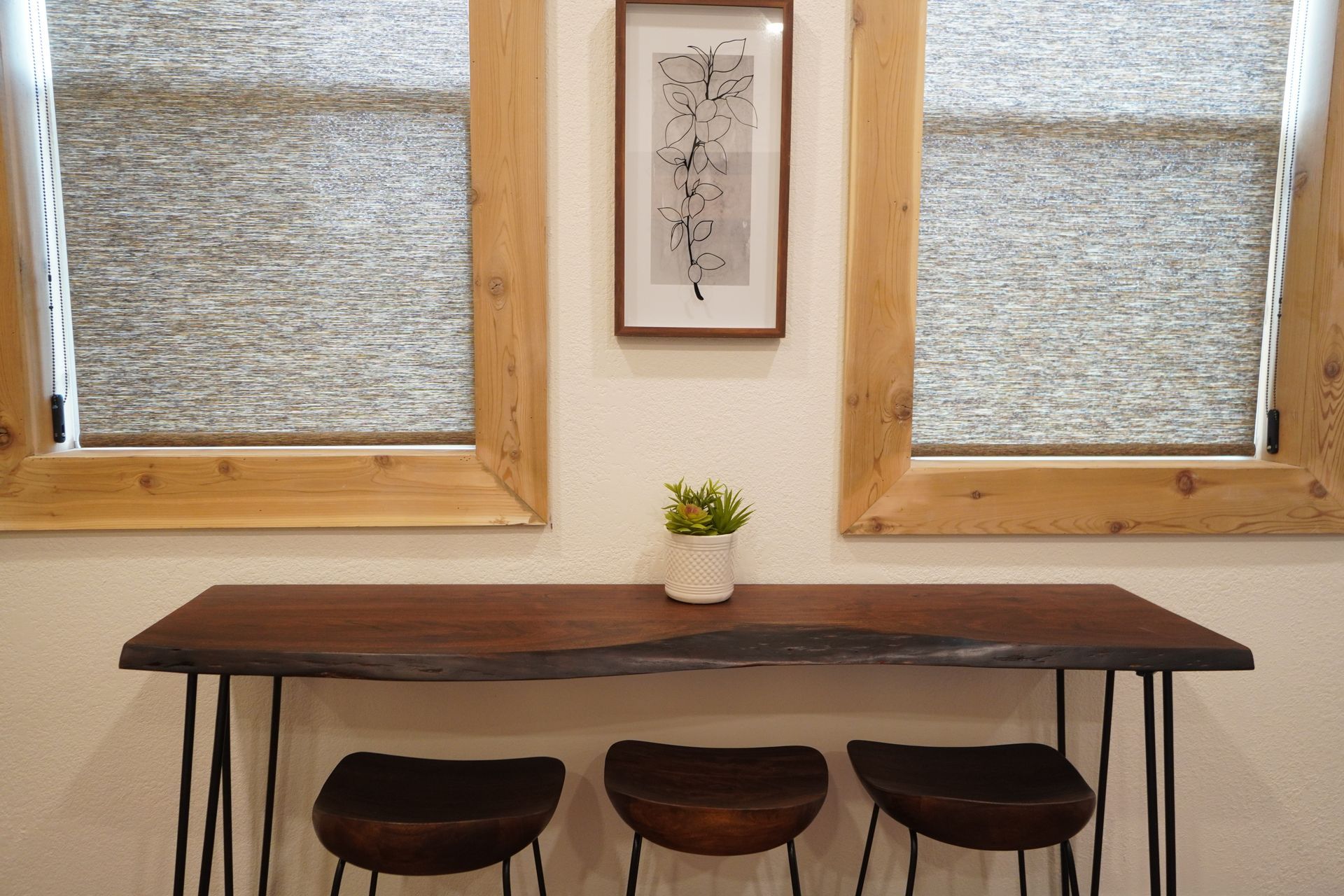 Wooden console table with three stools below windows with shades, framed art, and a small plant.