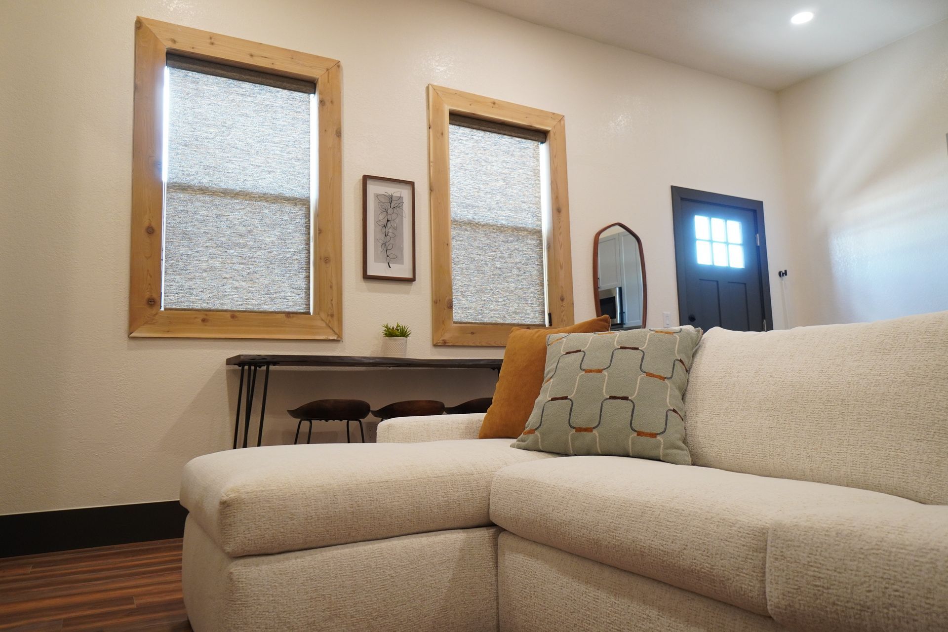 Cozy living room with a cream-colored sectional sofa, two windows with blinds, and a dark wooden door.