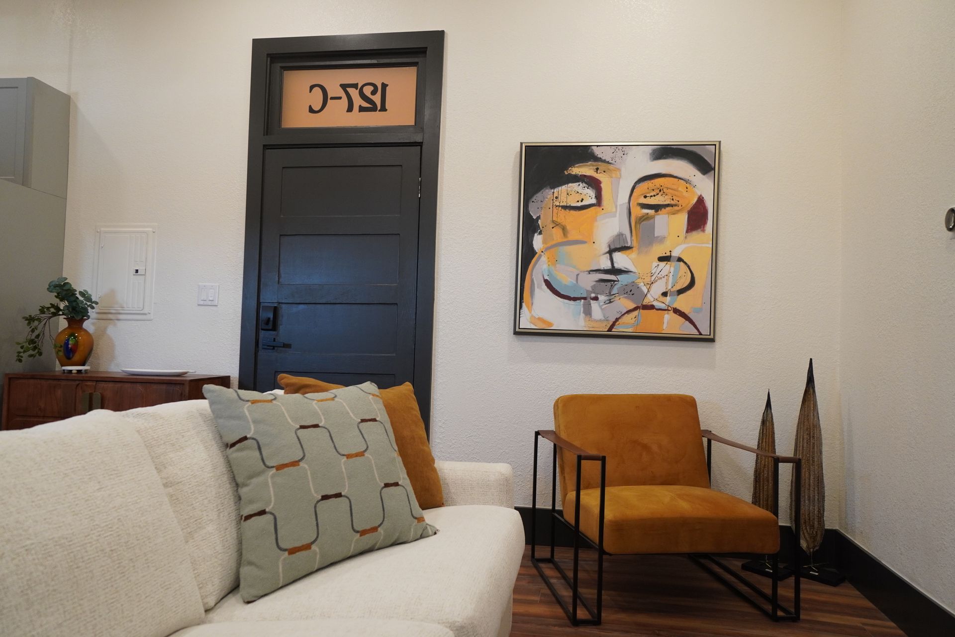 Cozy living area with a white sofa, orange chair, and abstract art above it. Black door with unit number.