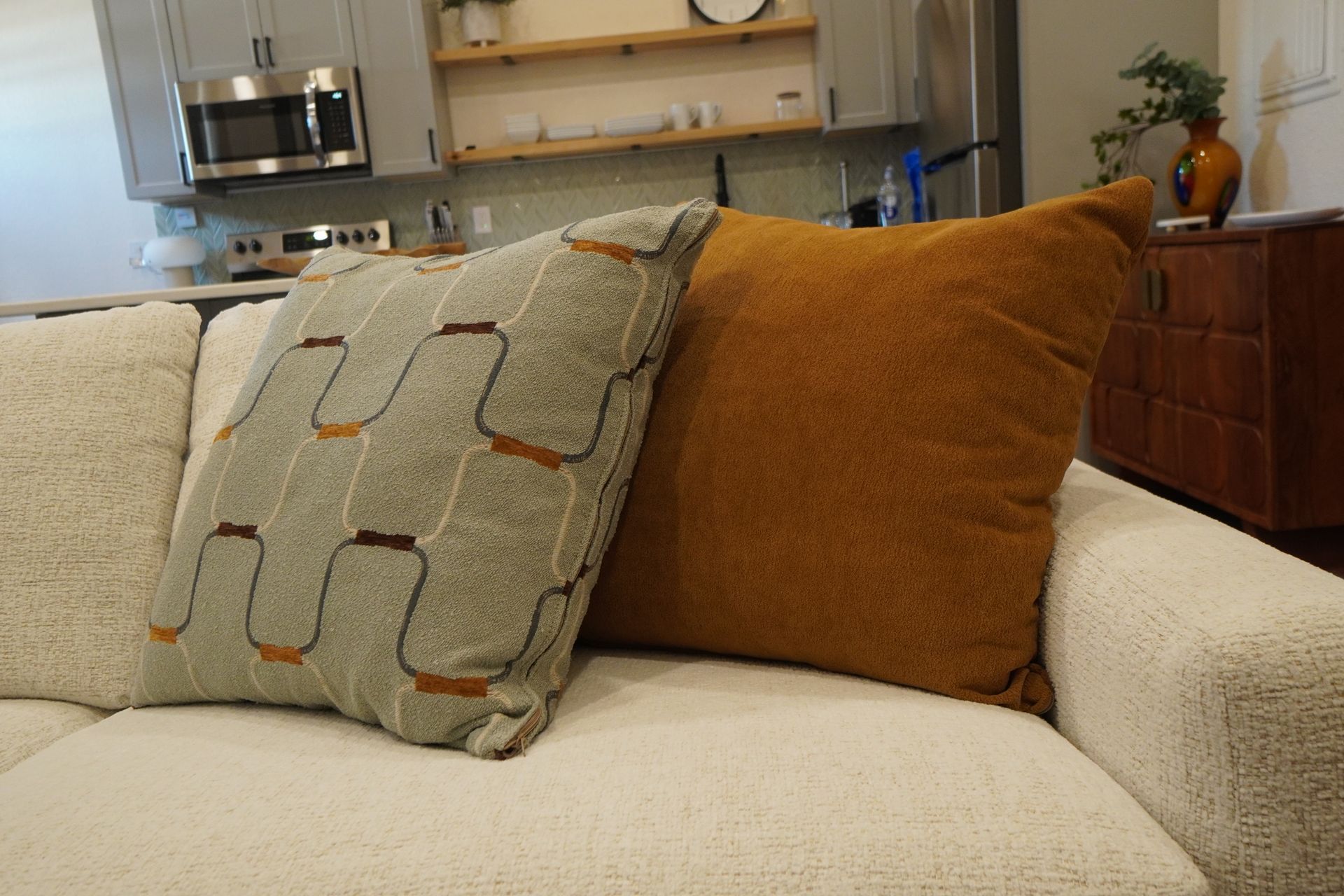 Two throw pillows, one mustard yellow, one patterned, on a white couch. Kitchen in background.