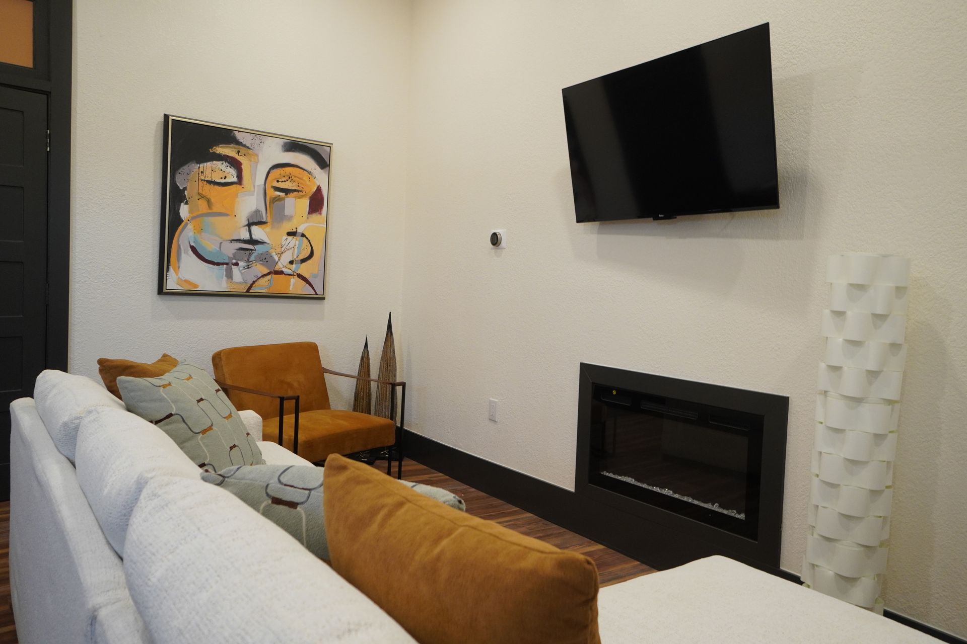 Living room with white sofa, fireplace, wall-mounted TV, orange chairs, and abstract art.